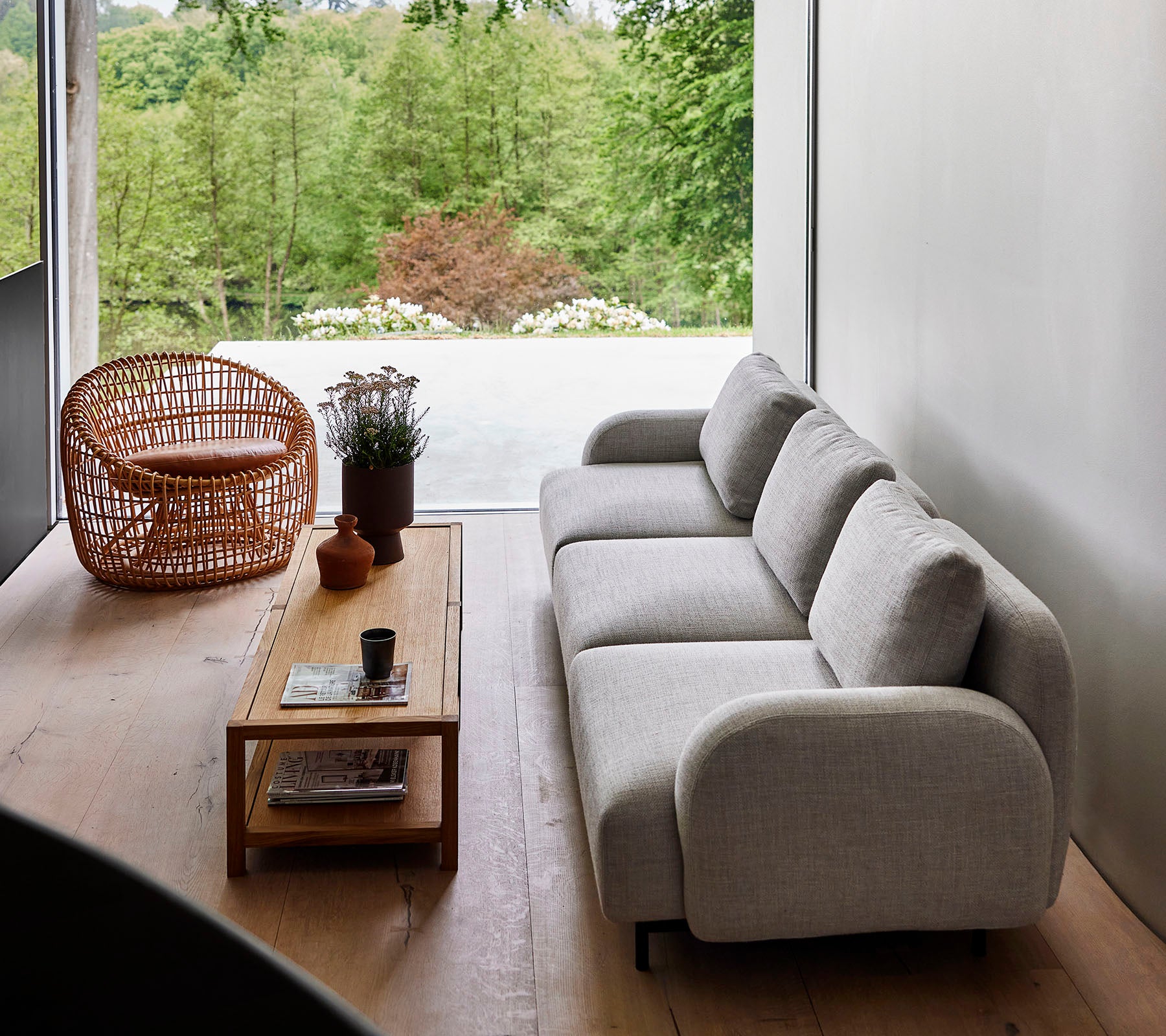 Moderne Sofa aus hellem Stoff, kombiniert mit einem Holztisch und einem Rattanstuhl, umgeben von Grünpflanzen. Natürliches Sonnenlicht verbessert die ruhige Atmosphäre.