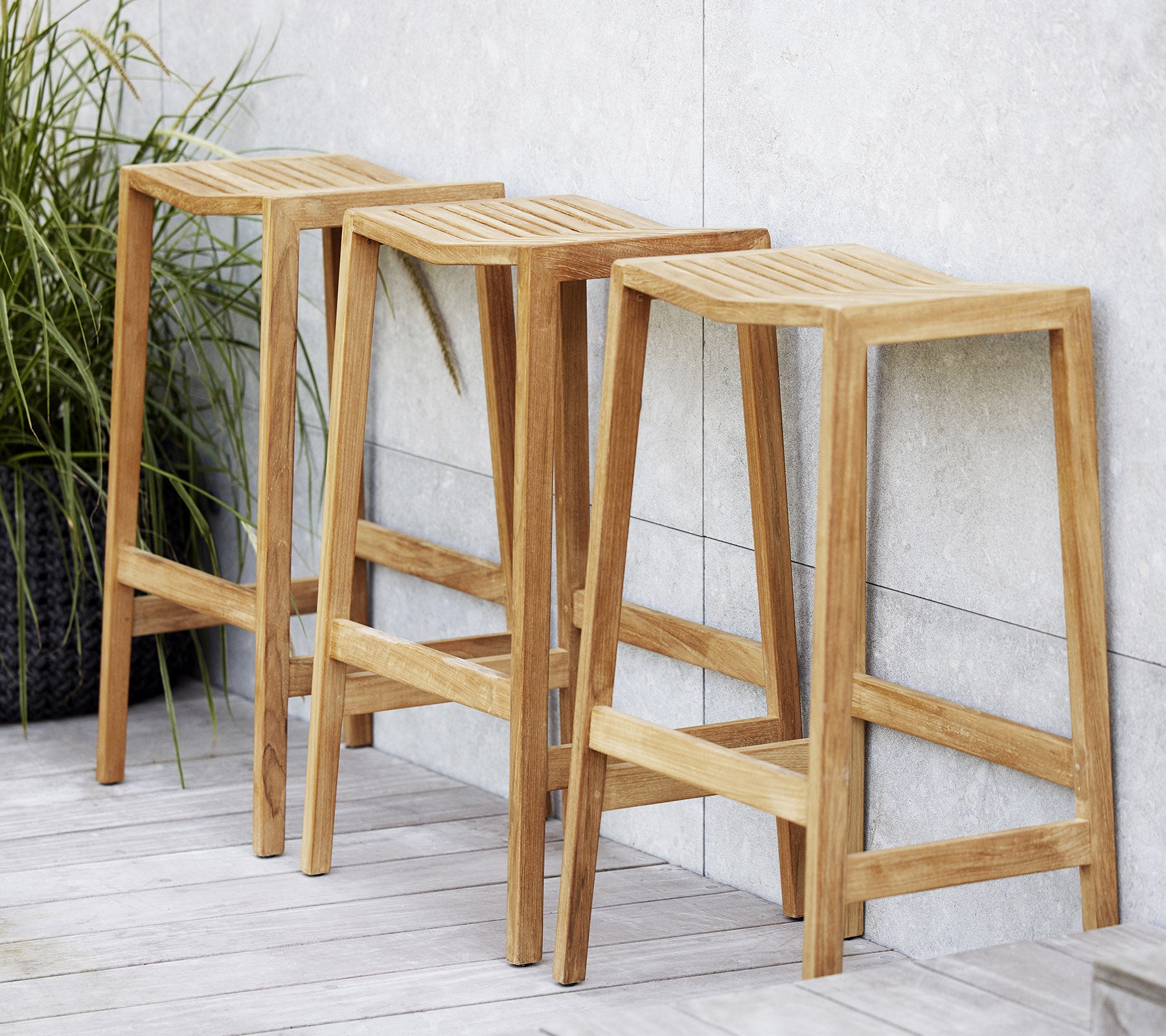 Drei Holz-Flip-Barhocker mit geschlitzten Sitzen, die ein minimalistisches Design und klare Linien aufweisen, neben einer dekorativen Pflanze positioniert.