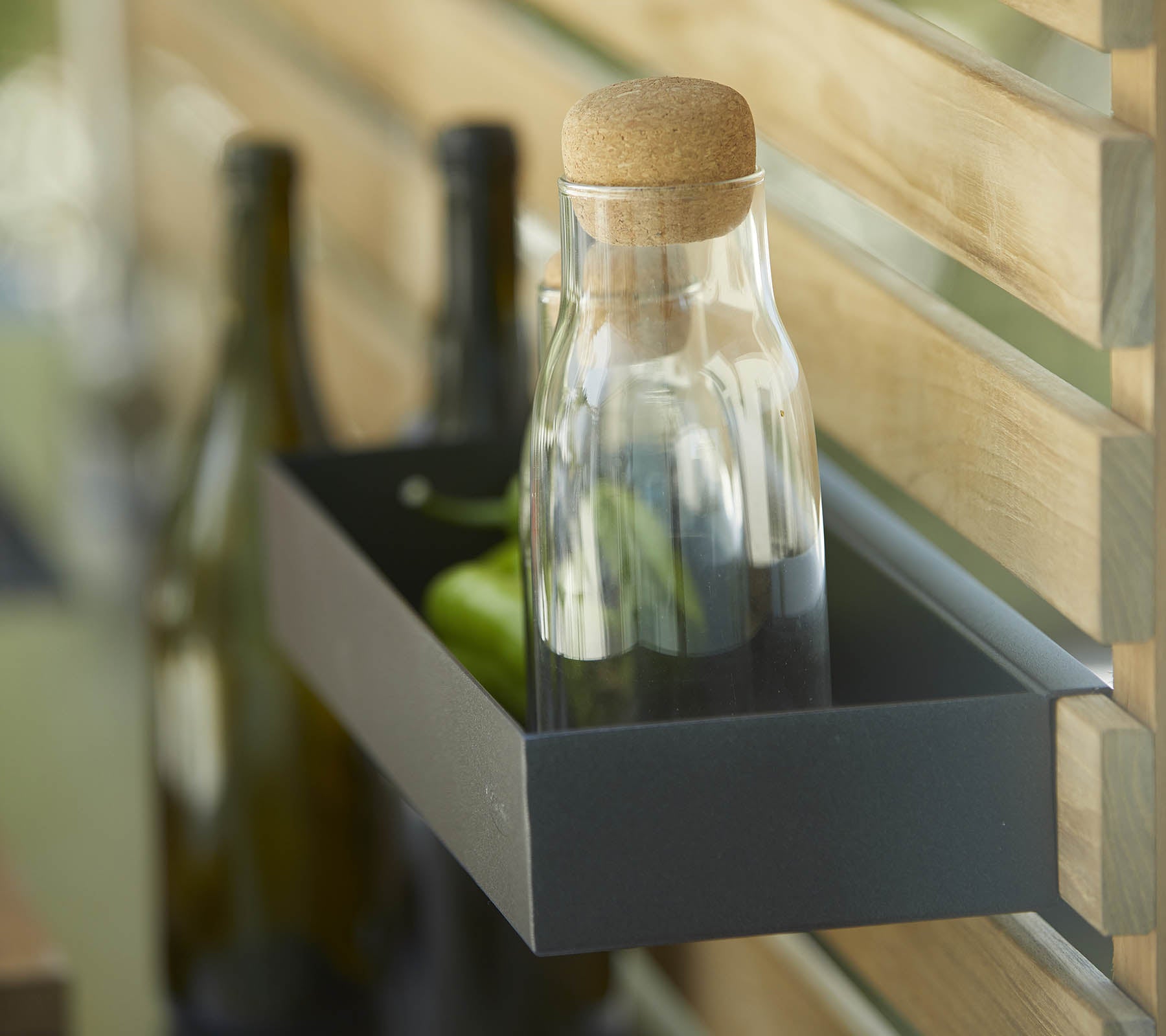Eine Glasflasche mit einem Korken steht auf einem Regal neben mehreren dunklen Flaschen, vor einem Holz-Hintergrund.
