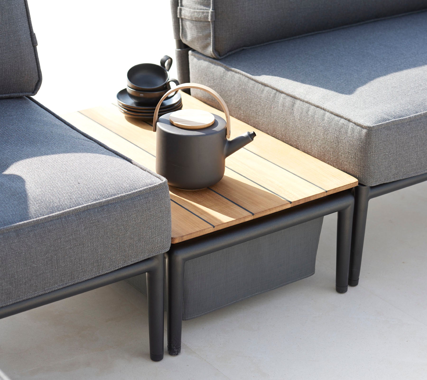 Zwei graue Sofas flankieren einen Holztisch, der mit einer schwarzen Teekanne und mehreren Tassen gedeckt ist, wodurch eine gemütliche Sitzecke entsteht.