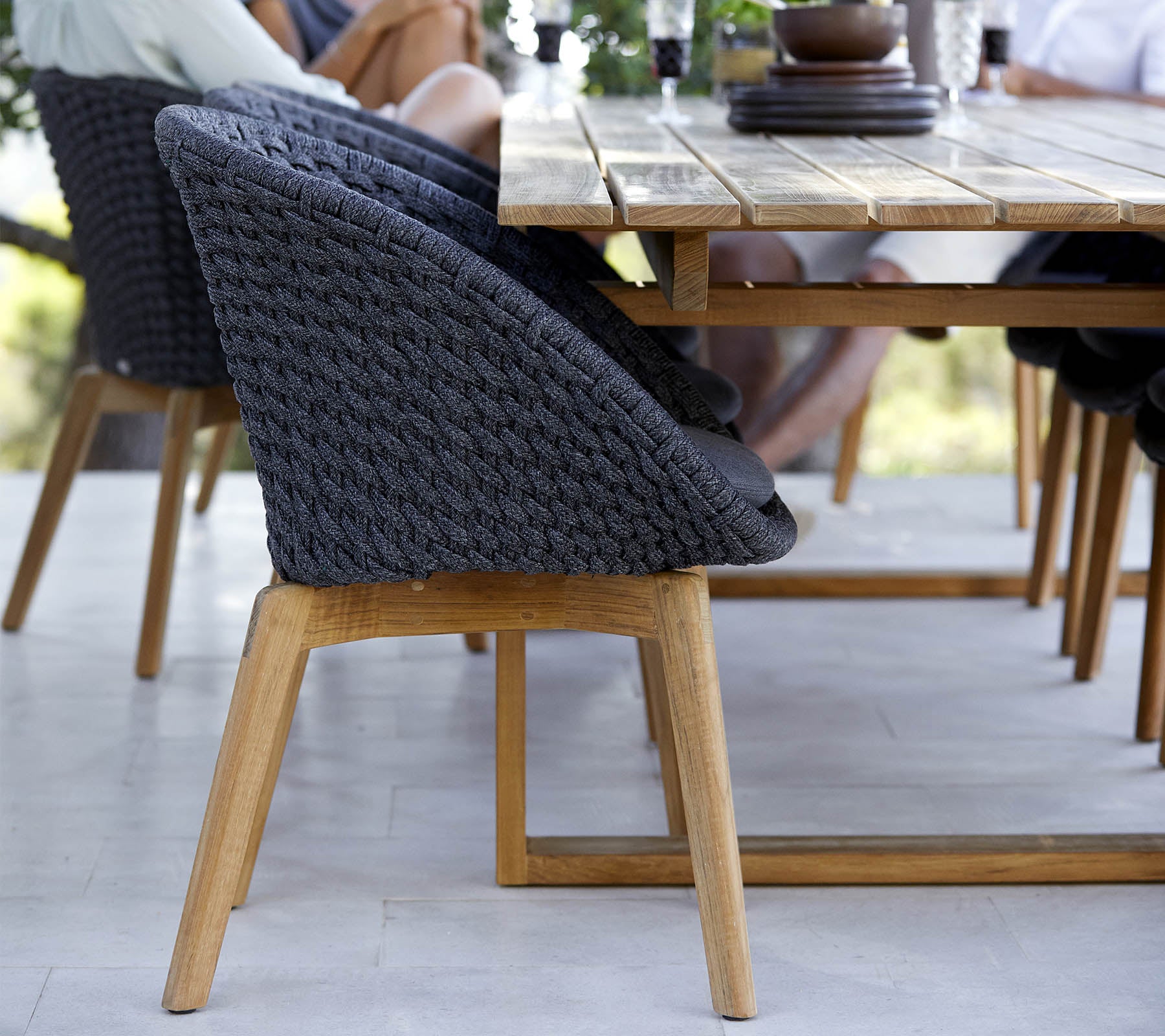 Ein stilvoller Stuhl mit einem geflochtenen Rücken und Beinen aus Holz, neben einem Holztisch platziert, mit Leuten in der Nähe, die beschäftigt sind.