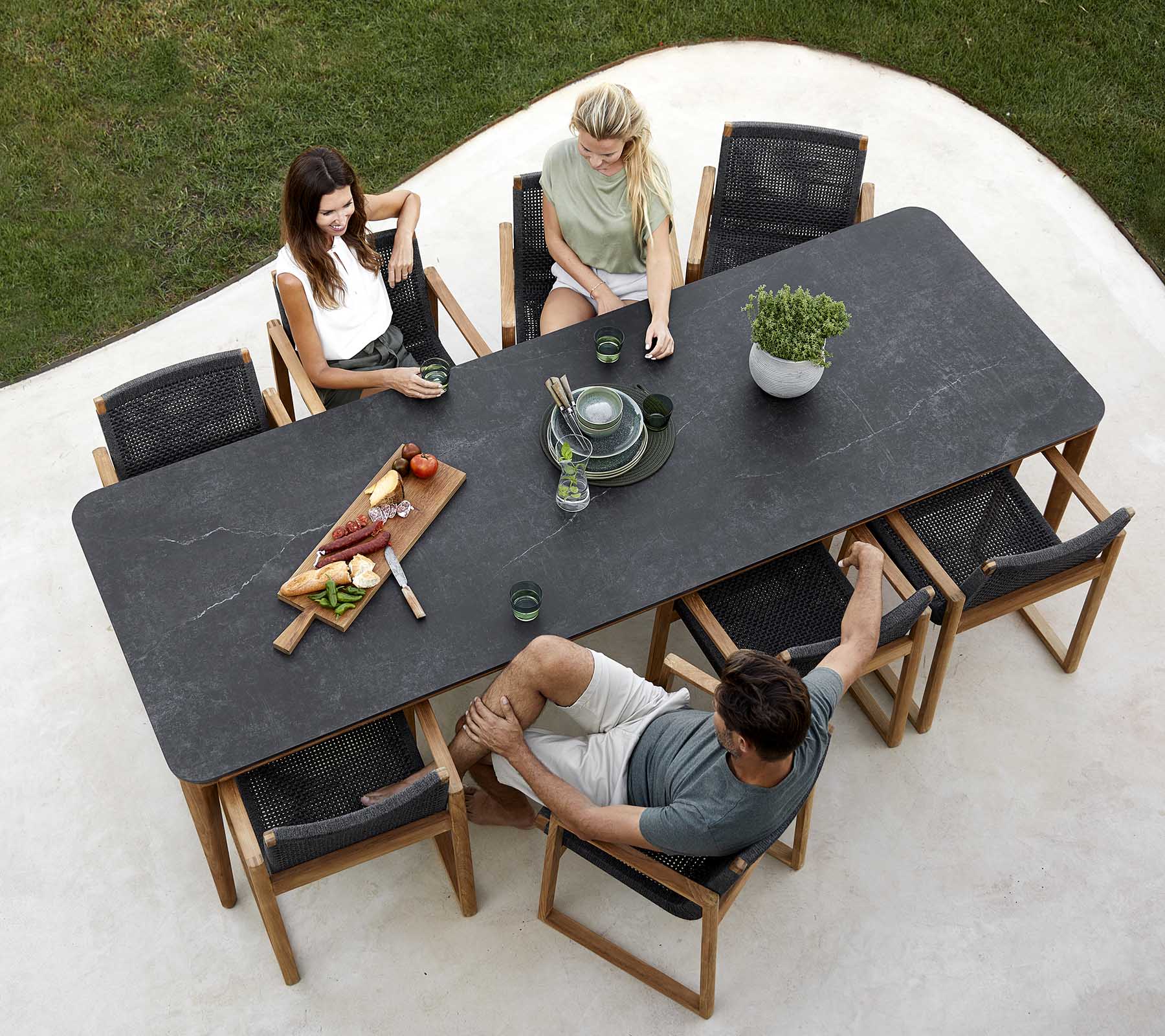 Eine Gruppe von Personen genießt eine Mahlzeit an einem großen Tisch mit Gerichten, Getränken und einem Pflanzenmittelstück. Informelle Ess-Atmosphäre.