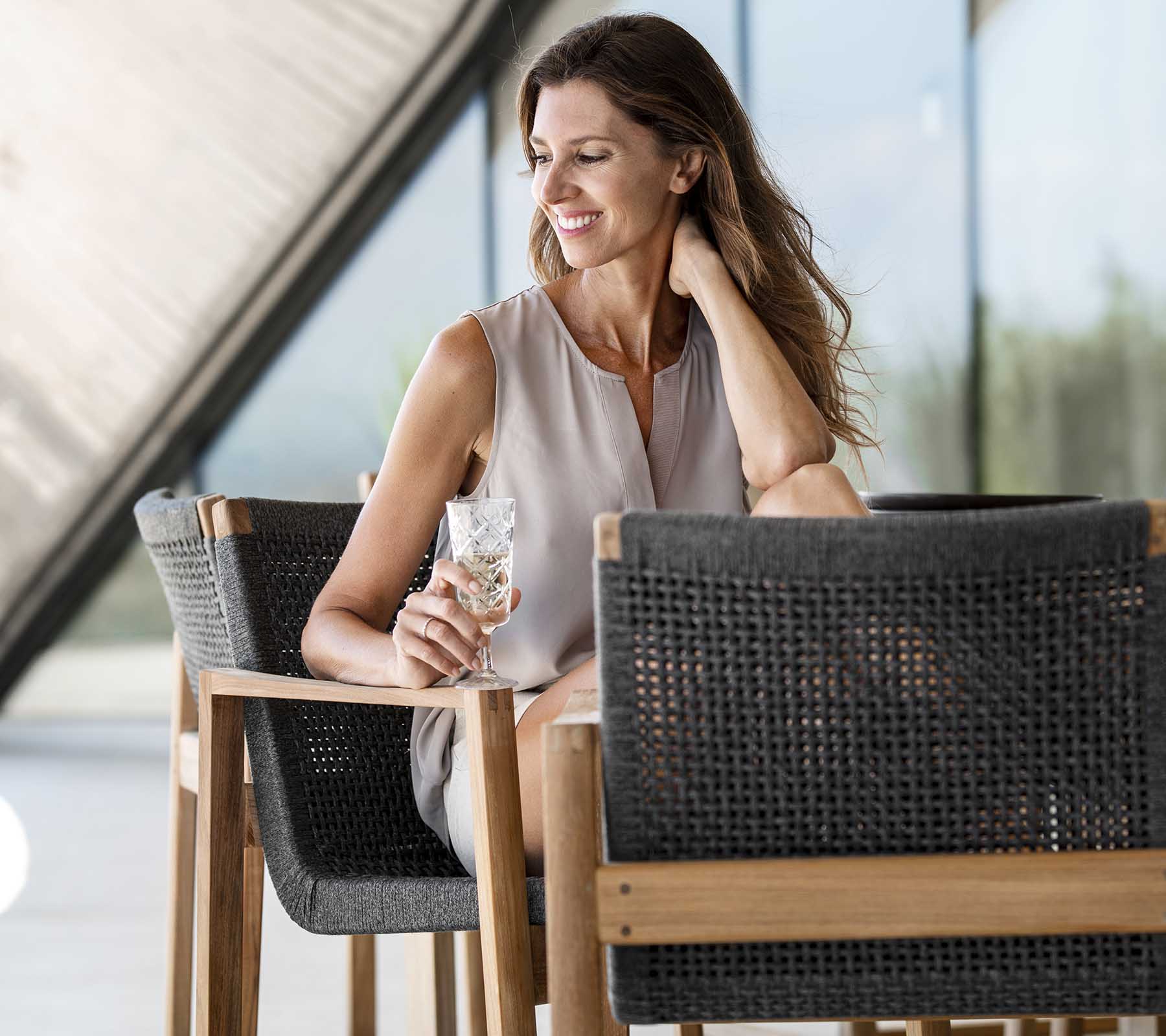 Lächelnde Frau hält ein Getränk, während sie auf einem stylischen Stuhl sitzt und eine entspannte, elegante Ausstrahlung hat. Helle und luftige Atmosphäre.