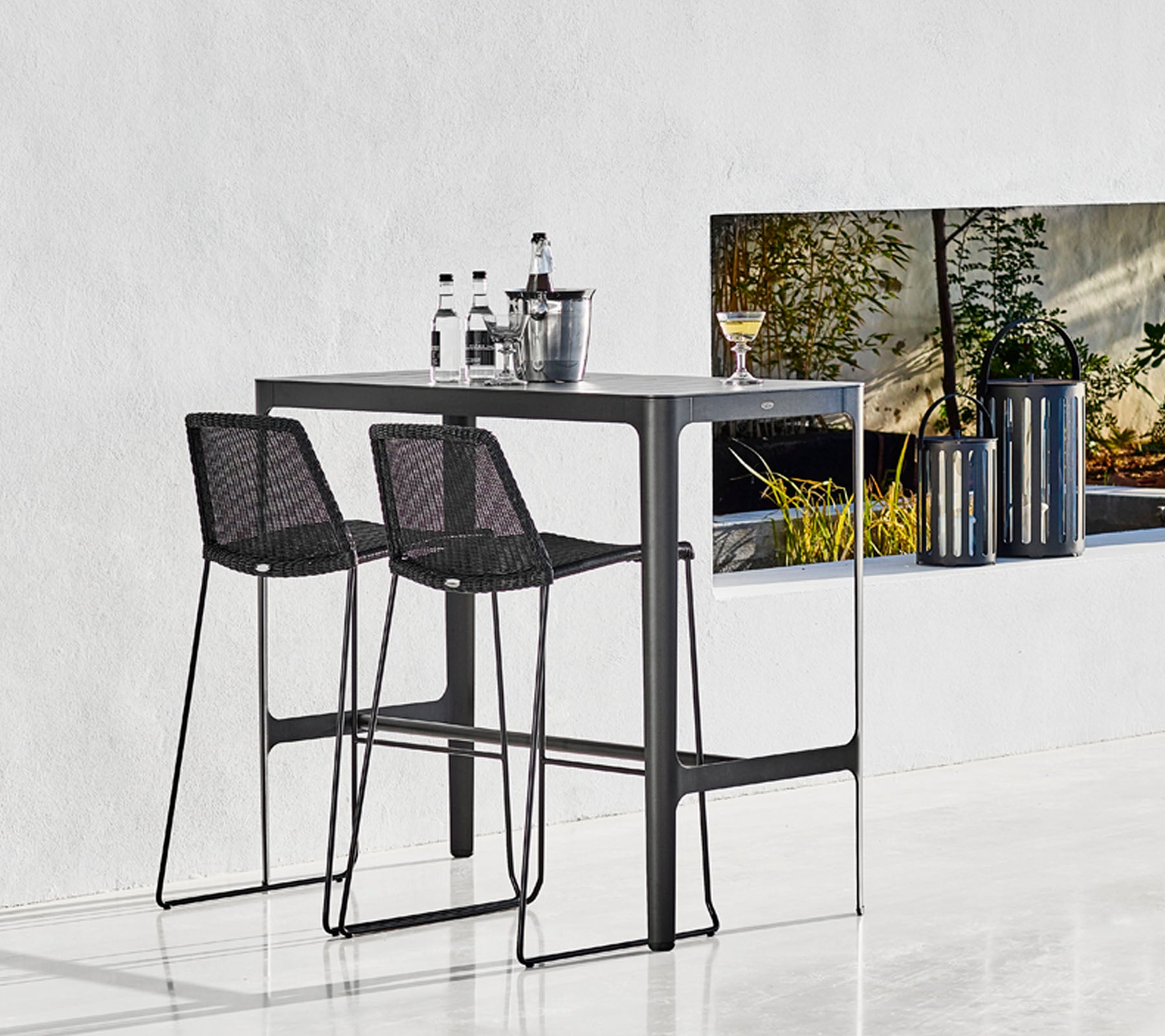 Eleganter Bartisch mit zwei schwarzen, geflochtenen Barhockern, begleitet von Glaswaren und einem stilvollen Eimer, vor einem minimalistischen Hintergrund.