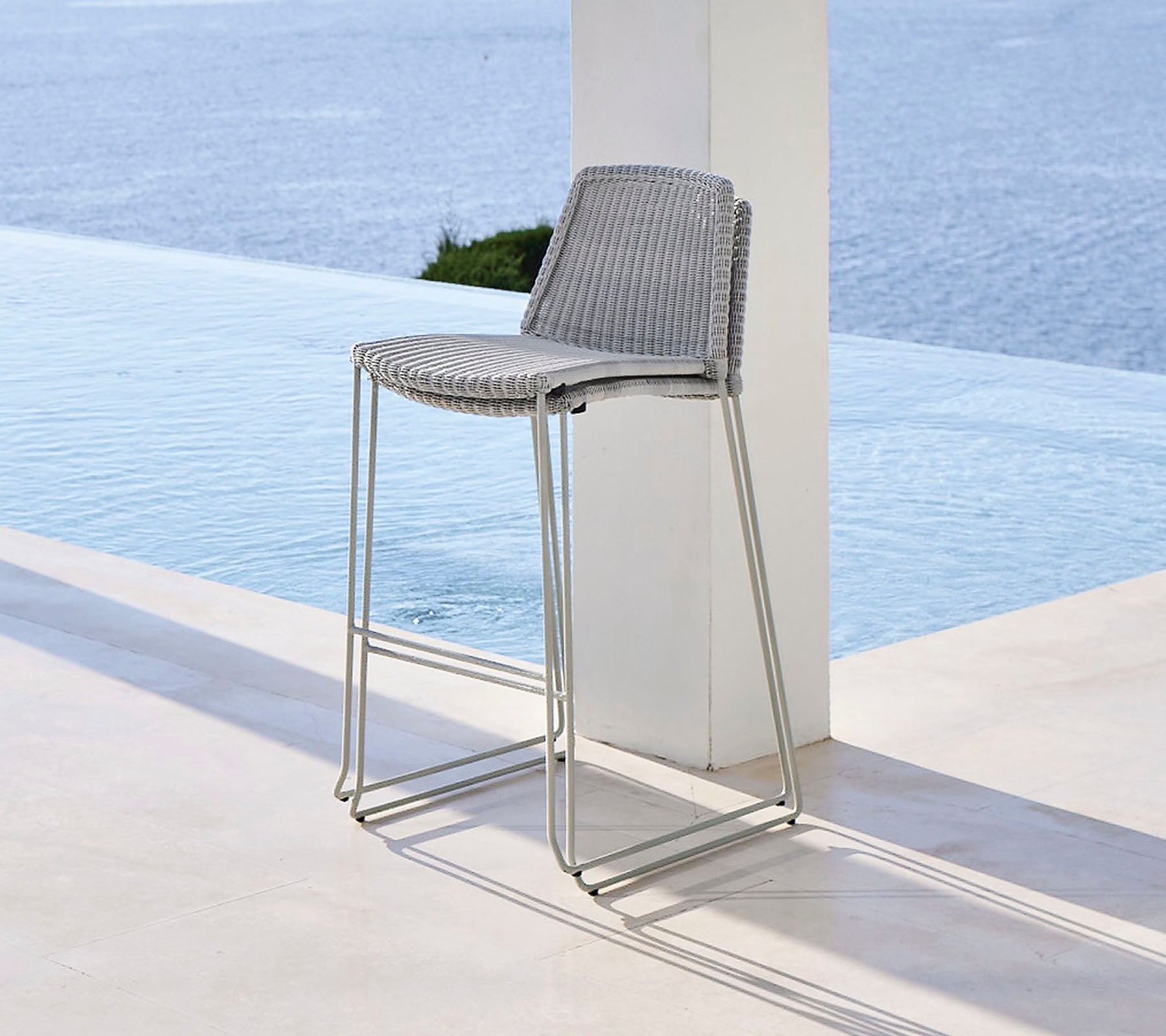 Stylischer Barhocker mit einem geflochtenen Sitz und schlanken Metallbeinen, positioniert in der Nähe eines ruhigen Wasserfeatures.