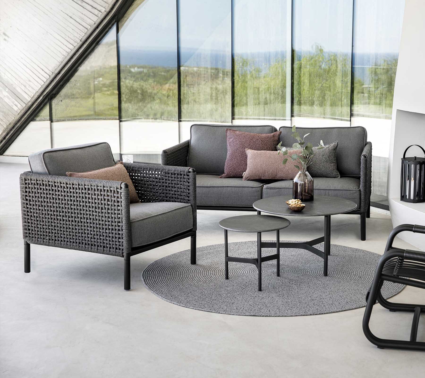 Stilvolle Sitzanordnung mit einem grauen Sofa und einem Sessel mit Kissen, begleitet von einem runden Tisch auf einem strukturierten Teppich.