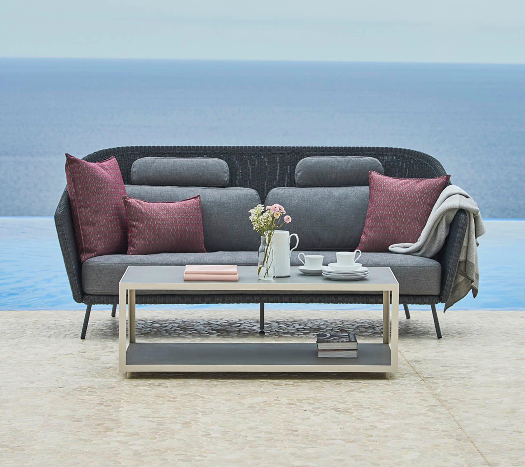 Stylische graue Couch mit dekorativen Kissen und einer gemütlichen Decke, ergänzt durch einen eleganten Couchtisch mit einer Teekanne und Büchern, vor einer ruhigen Landschaft.