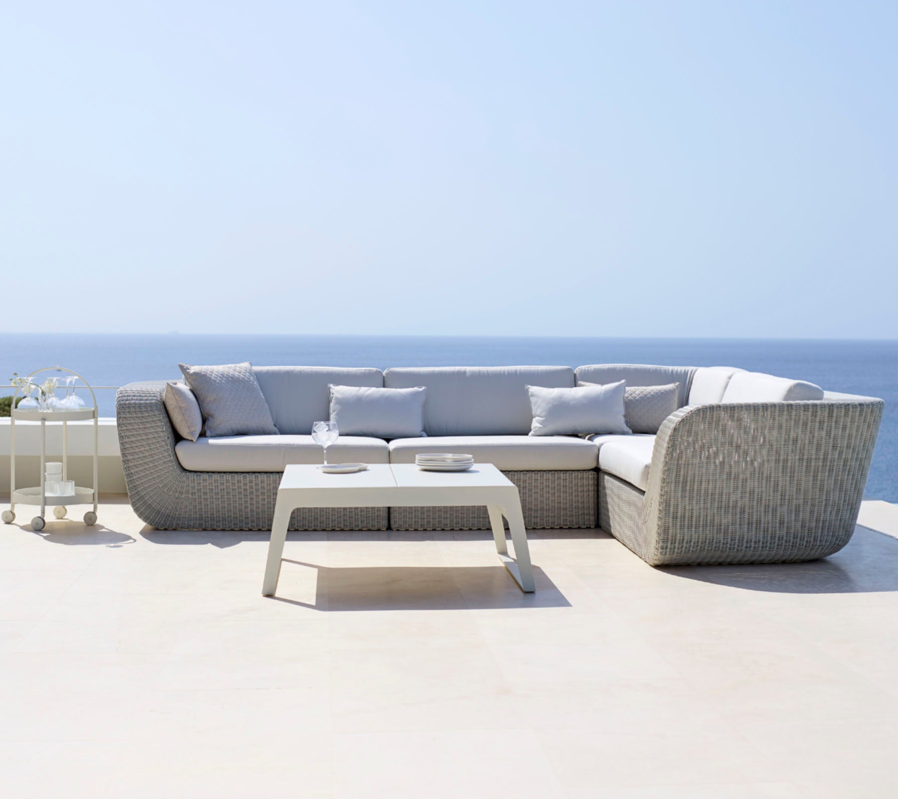 Modernes Ecksofa mit hellgrauen Kissen, kombiniert mit einem schlanken weißen Couchtisch, der vor einem ruhigen Ozeanhorizont steht.