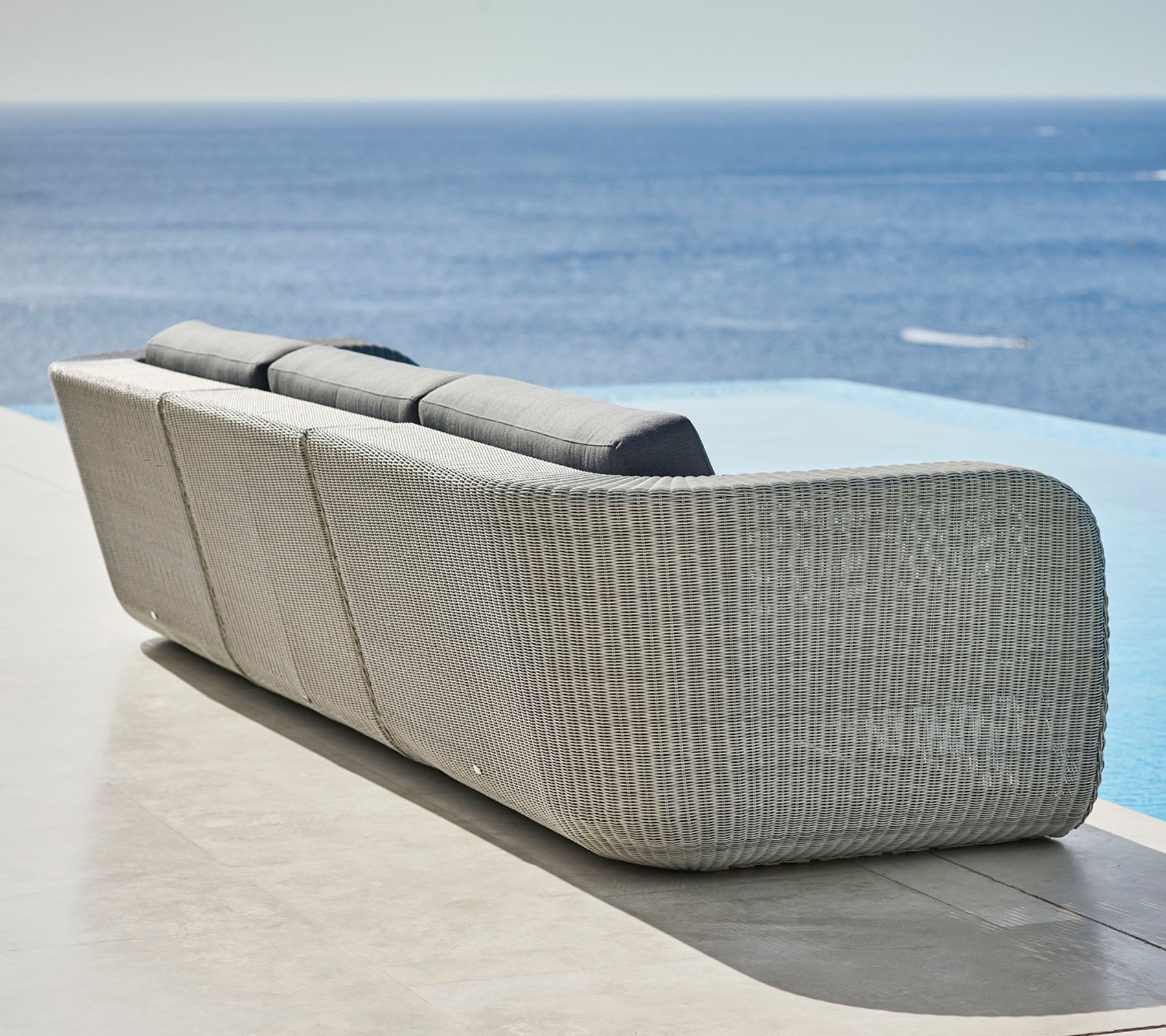 Stilvolles gewebtes Sofa mit geschwungenem Design und plüschigen Kissen mit Blick auf eine ruhige Meeresansicht. Ideal zur Entspannung und für Außenbereiche.