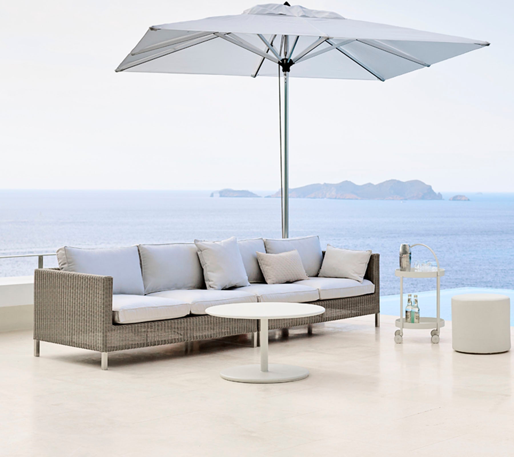 Stylisches Außenbereich mit einem hellgrauen Sofa, einem runden Tisch und einem großen Sonnenschirm mit Blick auf ein ruhiges Gewässer.