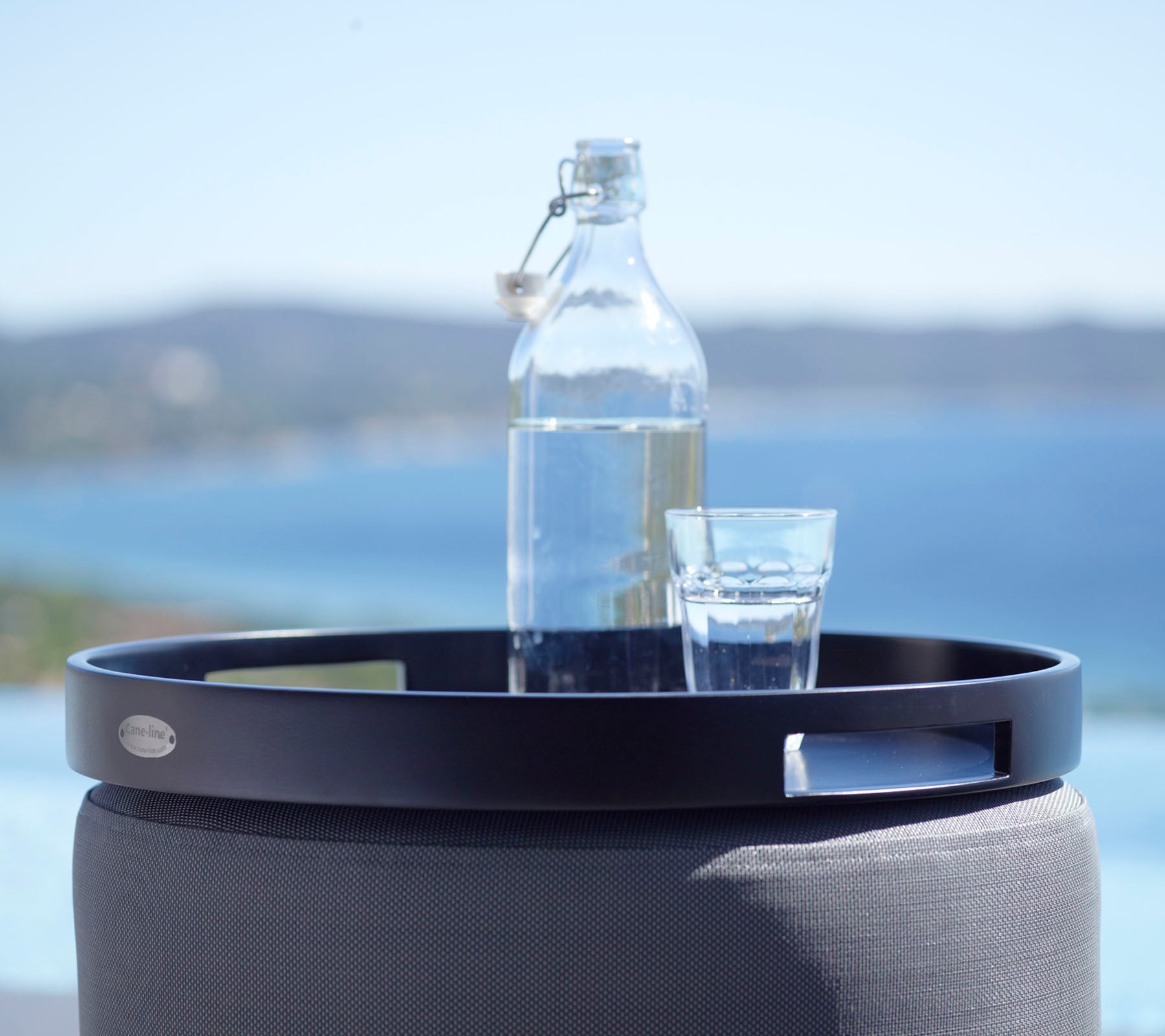 Ein schwarzes rundes Tablett hält ein Glas Wasser und eine Glasflasche, vor einer malerischen Kulisse aus dem Meer und fernen Hügeln.