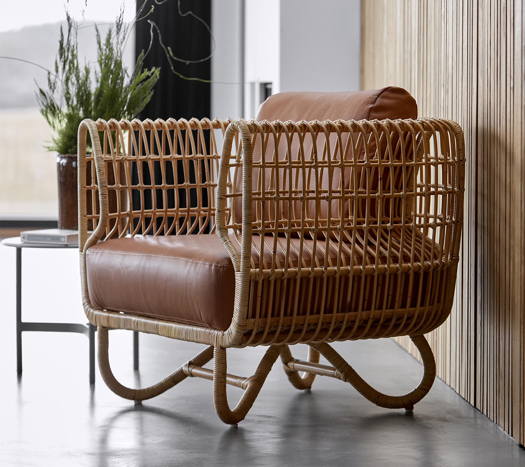 Stylisher Sessel mit einem geflochtenen Rattangehäuse und weichen braunen Lederkissen, perfekt für moderne Einrichtung.