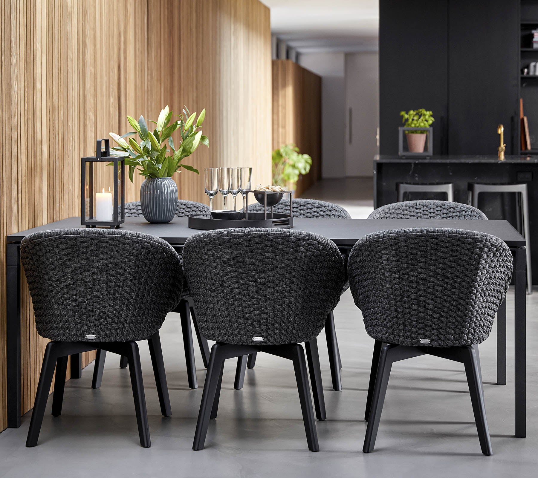 Ein modernes Esssetup mit einem schlanken schwarzen Tisch, umgeben von stilvollen geflochtenen Stühlen, ergänzt durch dekorative Pflanzen und Kerzen.