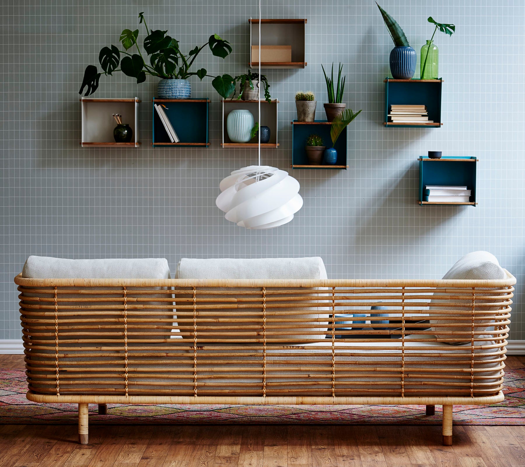 Ein stilvolles Rattansofa mit hellen Kissen vor einer strukturierten Wand, ergänzt durch wandmontierte Regale, die Pflanzen und dekorative Gegenstände展示.