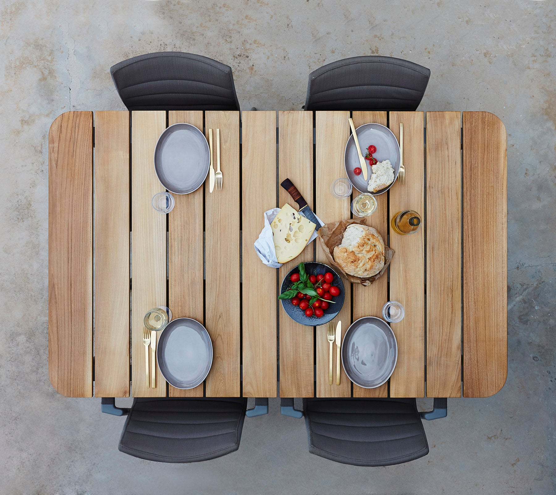 Ein Holztisch, gedeckt mit Tellern, Gläsern und einer Auswahl an Käse, Obst und Brot, umgeben von vier Stühlen.