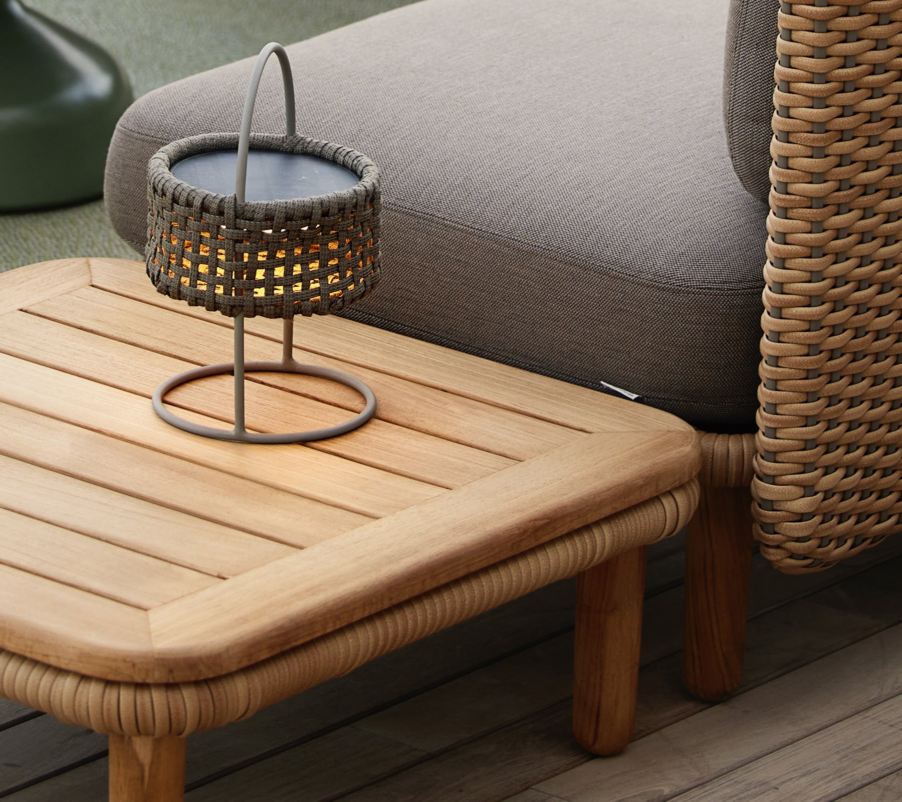Teak-Kaffeetisch mit einer lamellierten Oberfläche, begleitet von einer gewebten Lampe, die eine gemütliche und einladende Atmosphäre schafft.