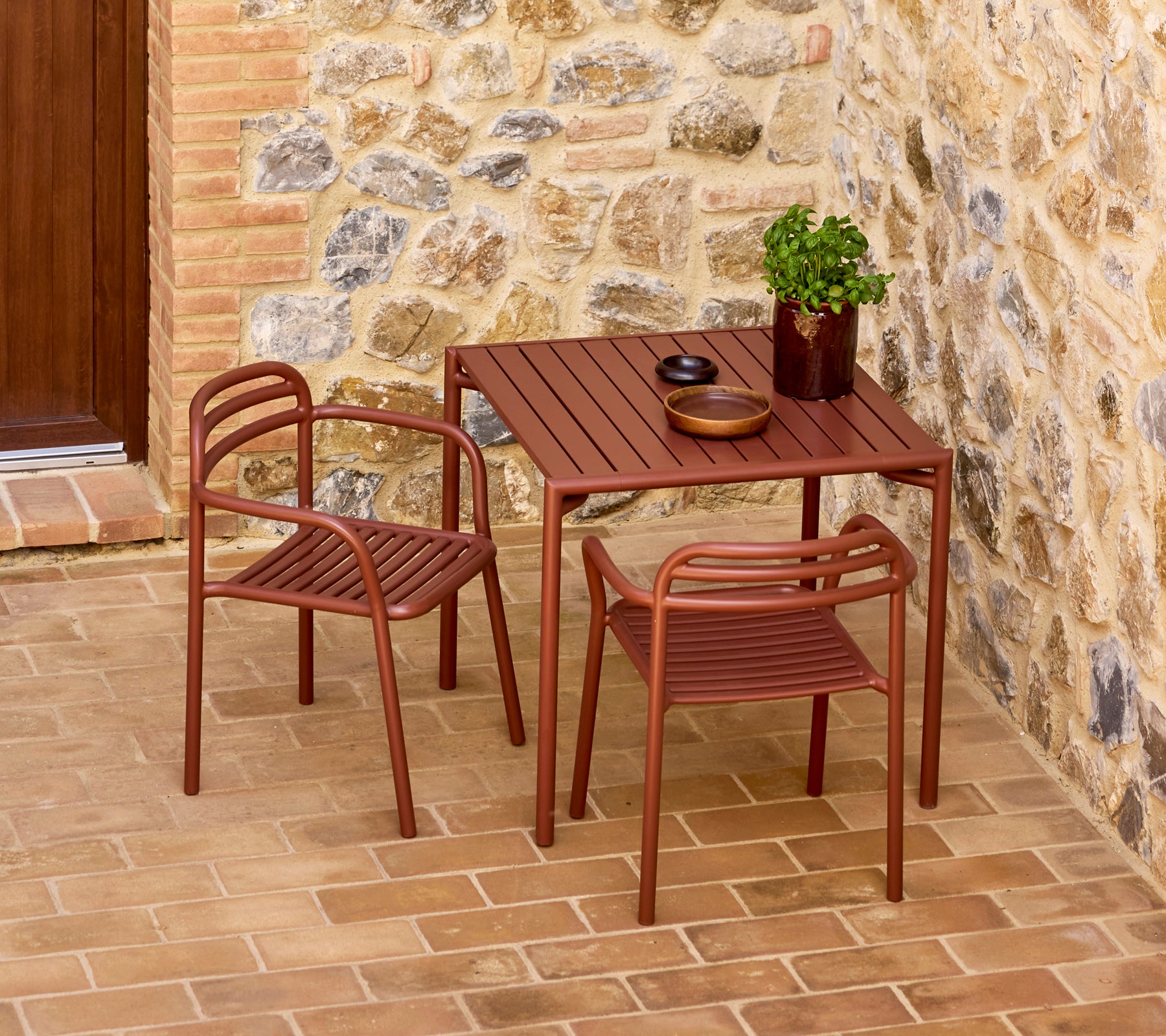 Eleganter scharlachroter Bliss-Tisch mit zwei passenden Stühlen, begleitet von einer dekorativen Pflanze und einfachem Geschirr vor einem Stein-Hintergrund.