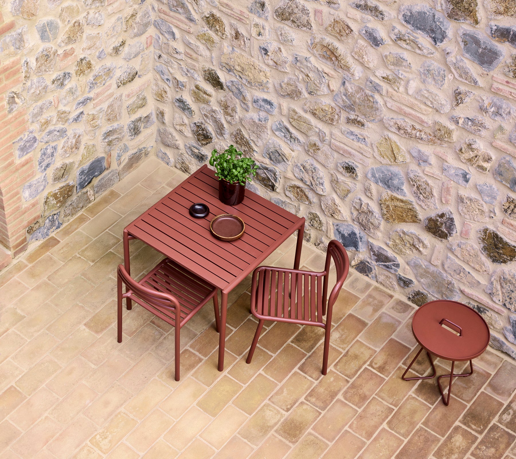 ein roter Café-Tisch mit zwei Stühlen, einem kleinen Pflanze und einem Teller, vor einer Steinmauer.