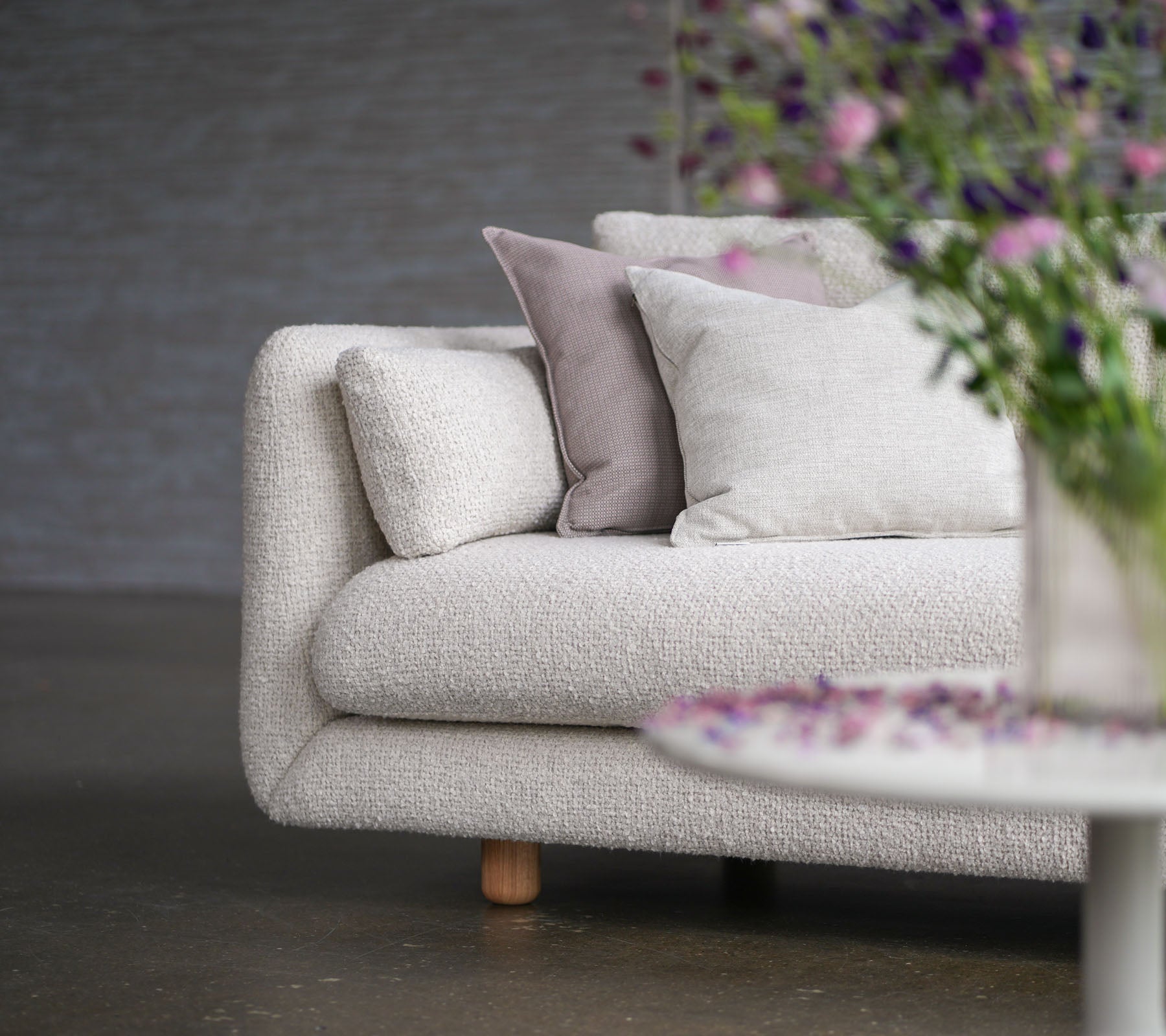 Weiches Sofa mit sandfarbenem Bezug, dekorativen Kissen, Teakholzbeinen und einer Vase mit Blumen, die Cane-line Flair und Eleganz präsentieren.