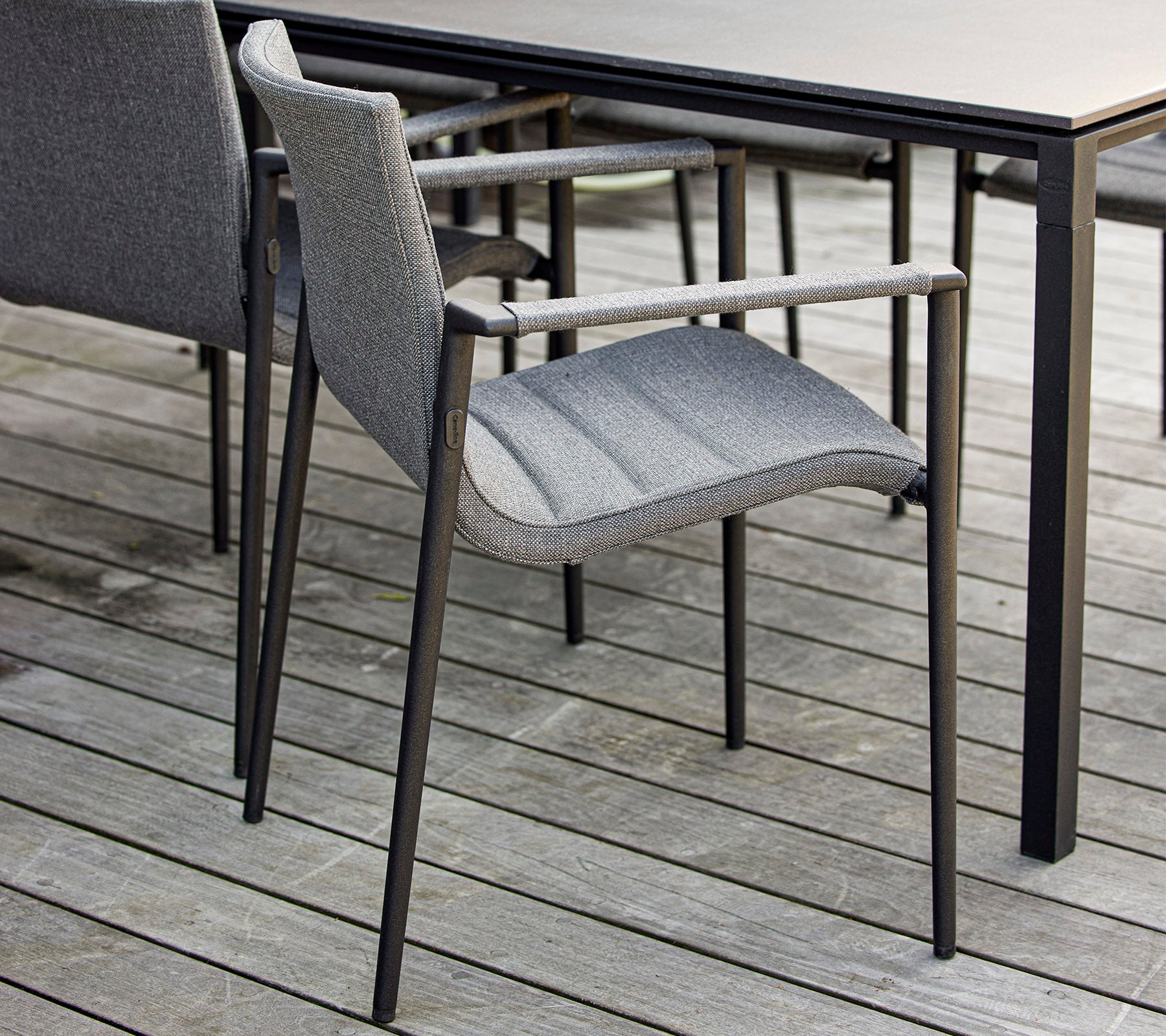 Stylischer grauer Stuhl mit Armlehnen, der neben einem eleganten schwarzen Tisch auf einer Holzoberfläche steht. Ideal für moderne Außenbereiche.
