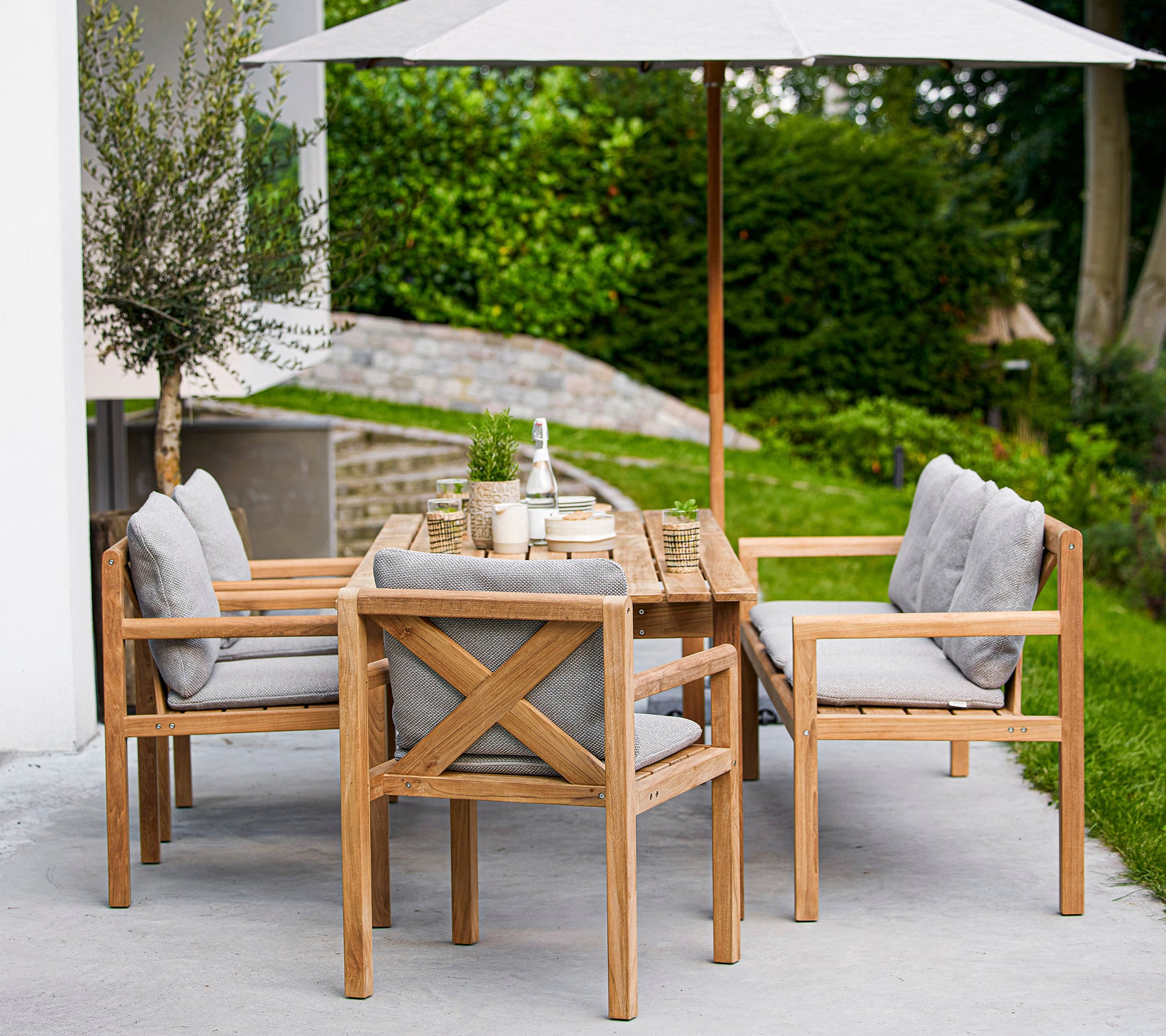 Holztischset mit einem rechteckigen Tisch und gepolsterten Stühlen, mit einem großen Sonnenschirm, der Schatten spendet. Grünflächen sind im Hintergrund sichtbar.