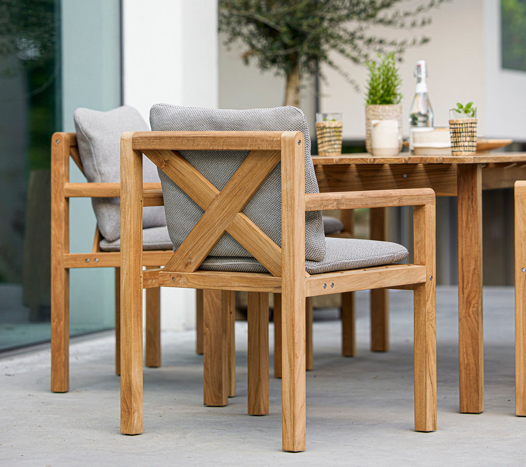 Holzstühle mit grauen Kissen und einem passenden Tisch, begleitet von dekorativen Pflanzen und einer Flasche, ideal für das Essen im Freien.