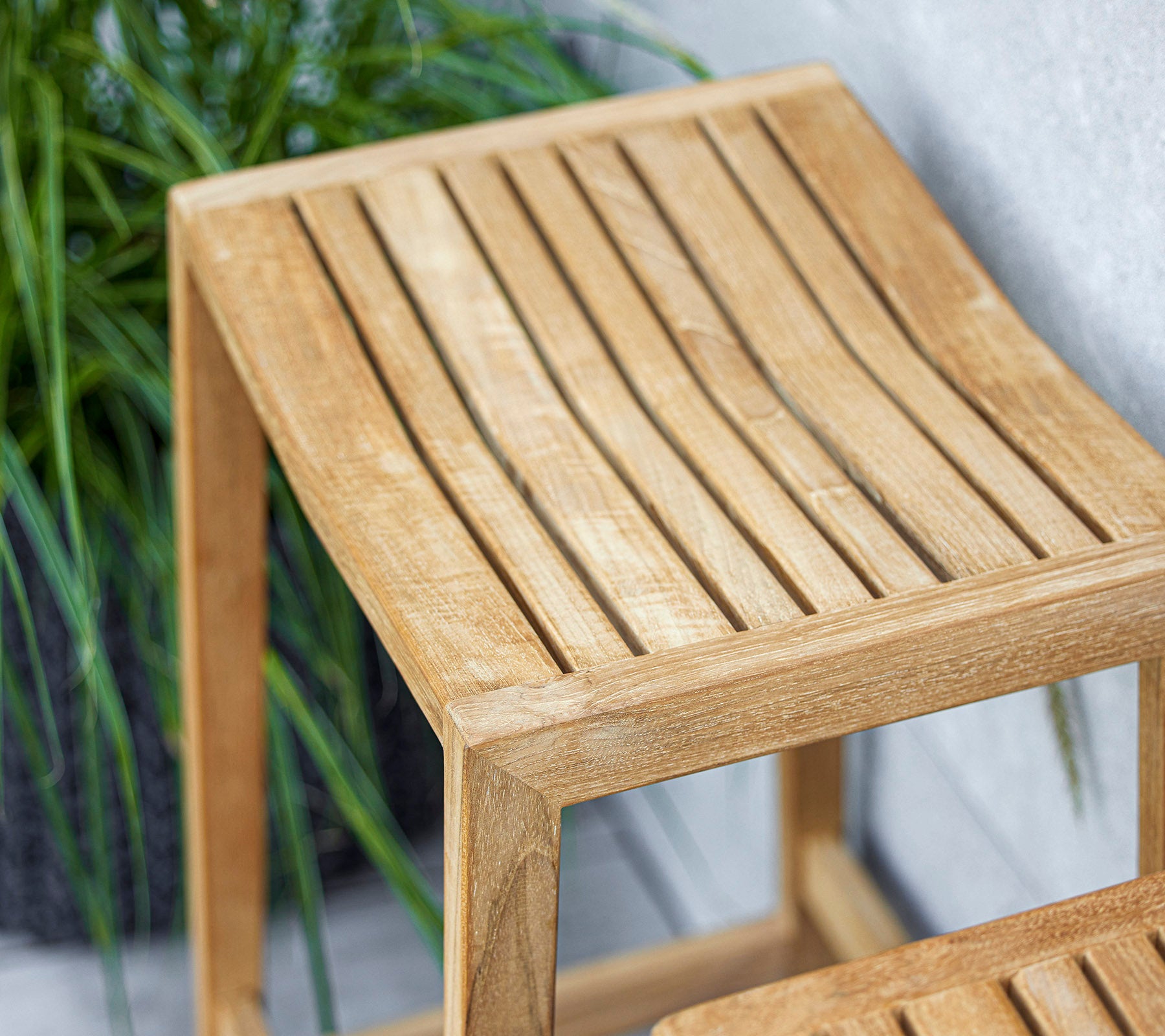 Ein schlanker Holztisch mit einem lamellenförmigen Oberdesign, das mit einer hellen Oberfläche die umgebende Grünpflanzen ergänzt.