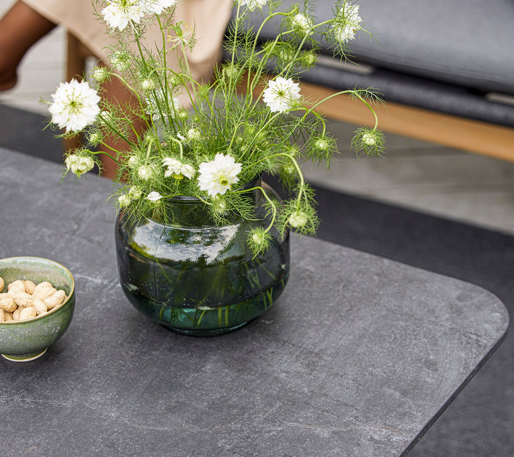 Ein modernes Tisch-Set mit einer runden Glasvase mit weißen Blumen und einer kleinen Schüssel Nüssen. Die Oberfläche ist glatt und strukturiert.