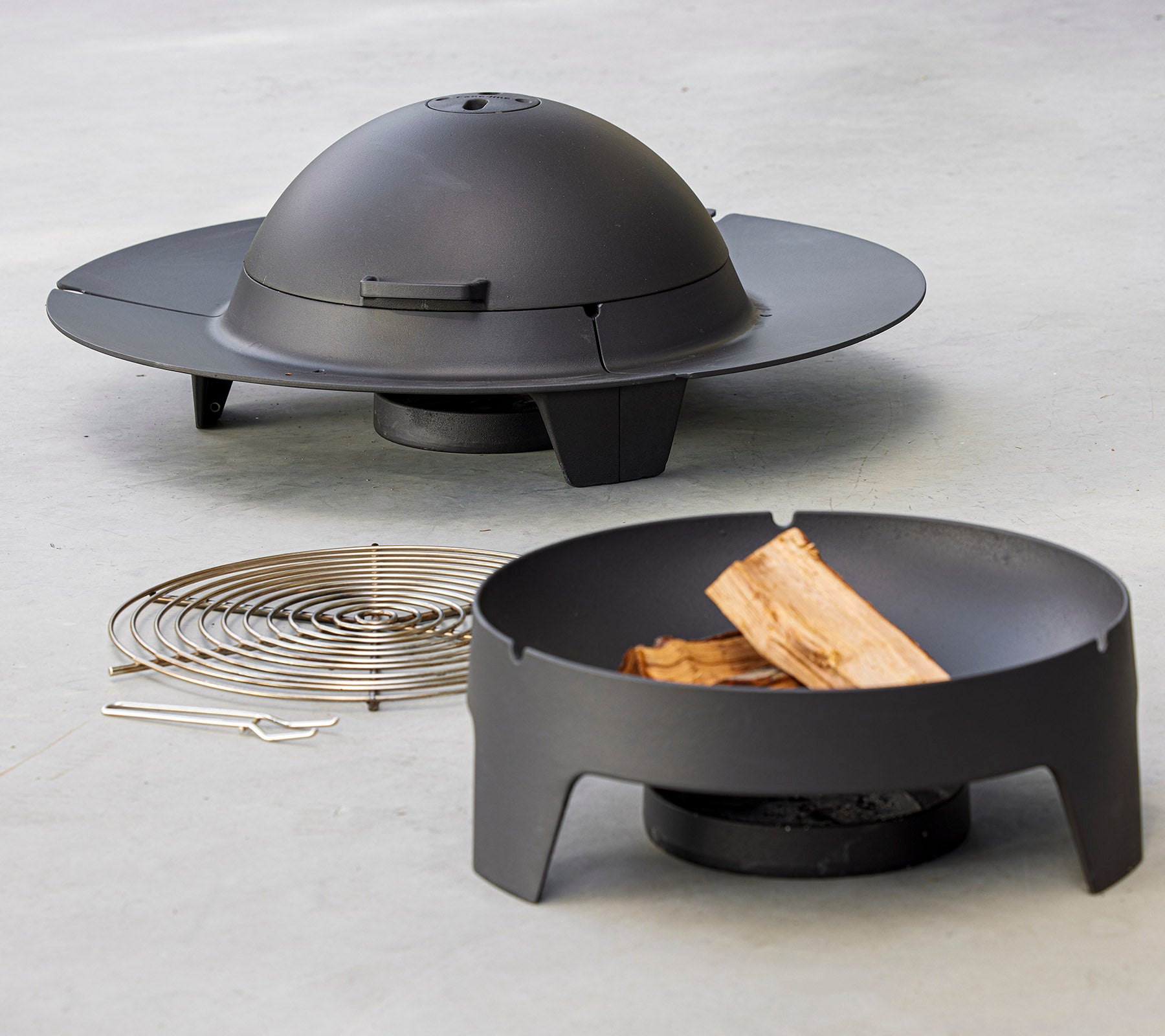 Ein modernes Feuerset mit einem schlanken, kreisförmigen Design und einem Kuppeldeckel, begleitet von einem Grillroster und Holzstücken.