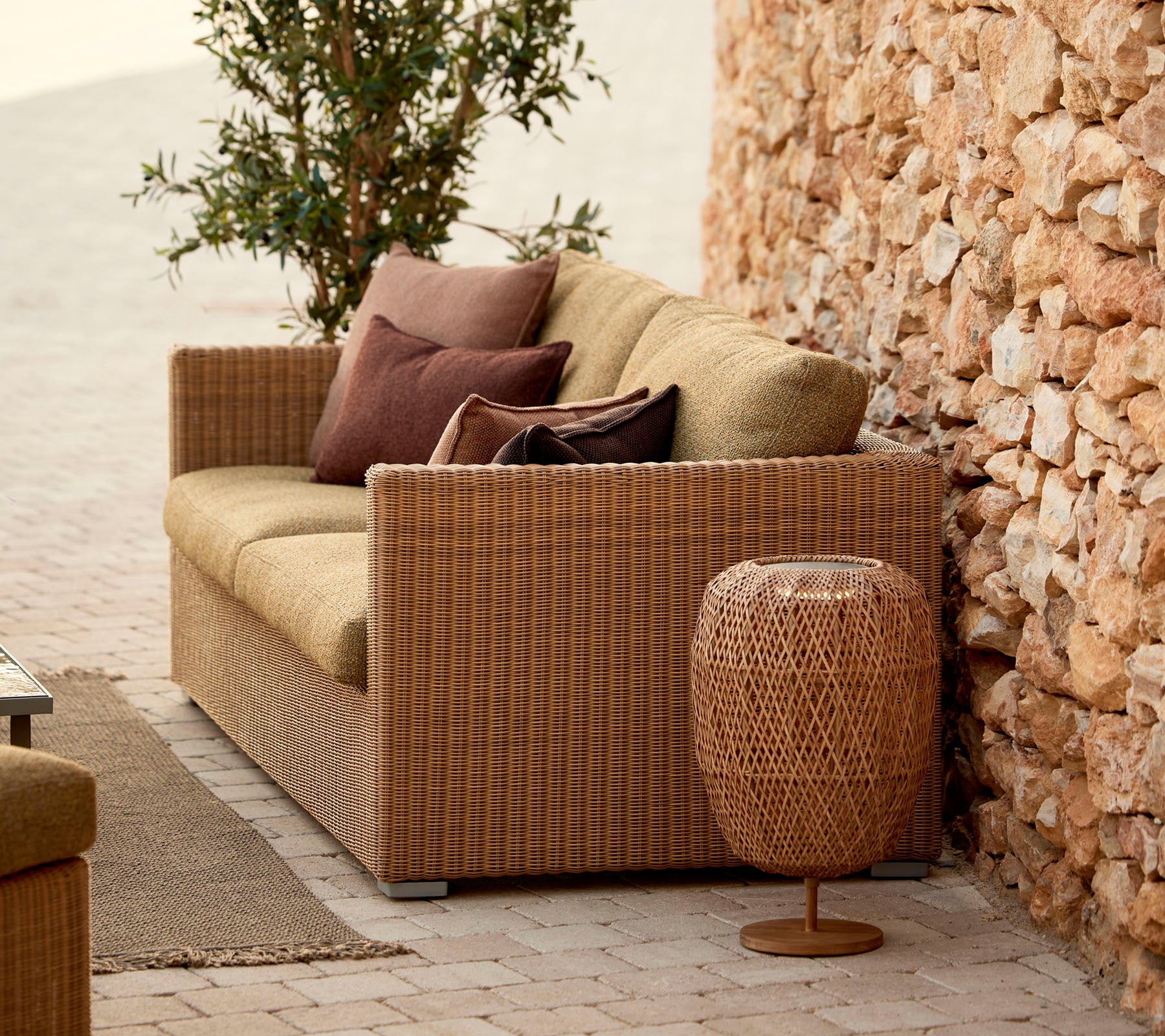 Eine gemütliche Rattansofa mit Kissen, ergänzt durch einen stilvollen geflochtenen Beistelltisch, vor einer Steinmauer und üppigem Grün.