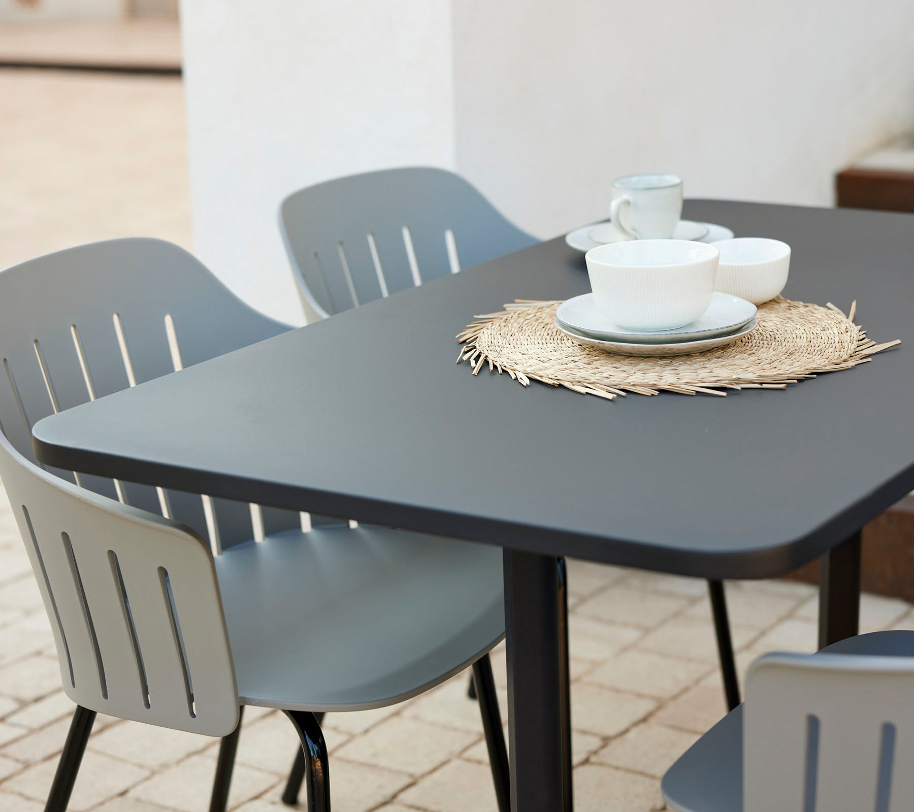 Modernes Cafétisch-Set mit einer schwarzen Tischplatte, umgeben von grauen Stühlen, mit Tellern und Tassen auf einem Untersetzer aus Stroh.