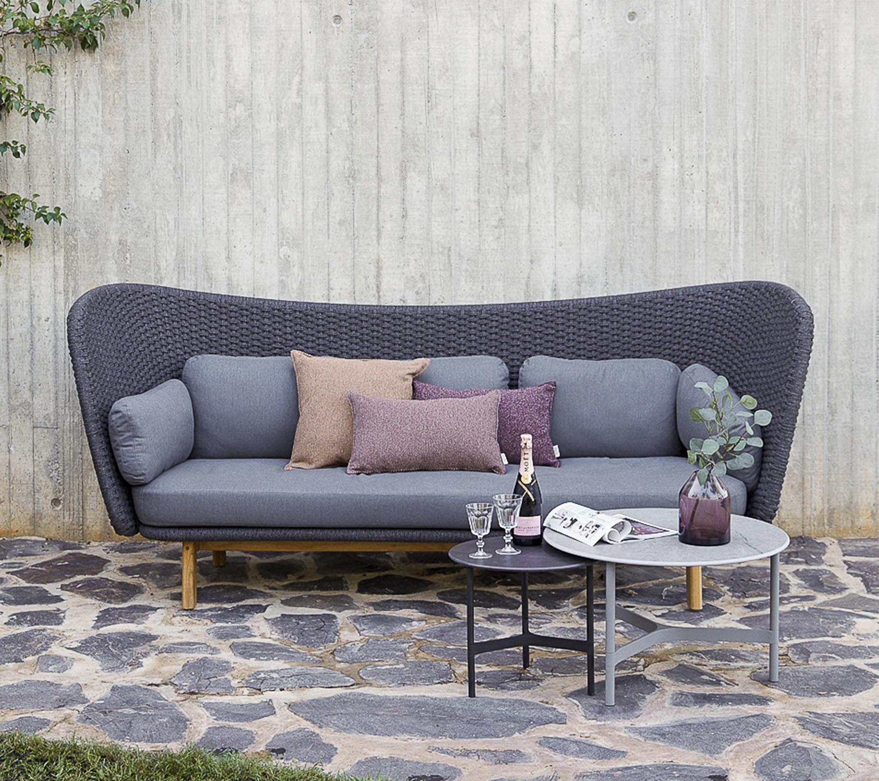 Ein modernes Sofa in Grau mit dekorativen Kissen, begleitet von einem kleinen Tisch mit Getränken und einer Vase, vor einer strukturierten Wand platziert.