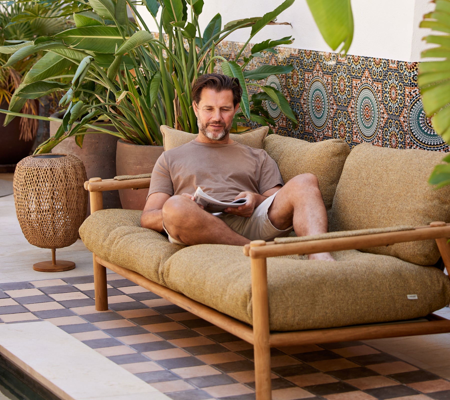 Ein Mann entspannt sich auf einem bequemen Sofa, liest eine Zeitschrift, umgeben von üppigem Grün und dekorativen Mustern.