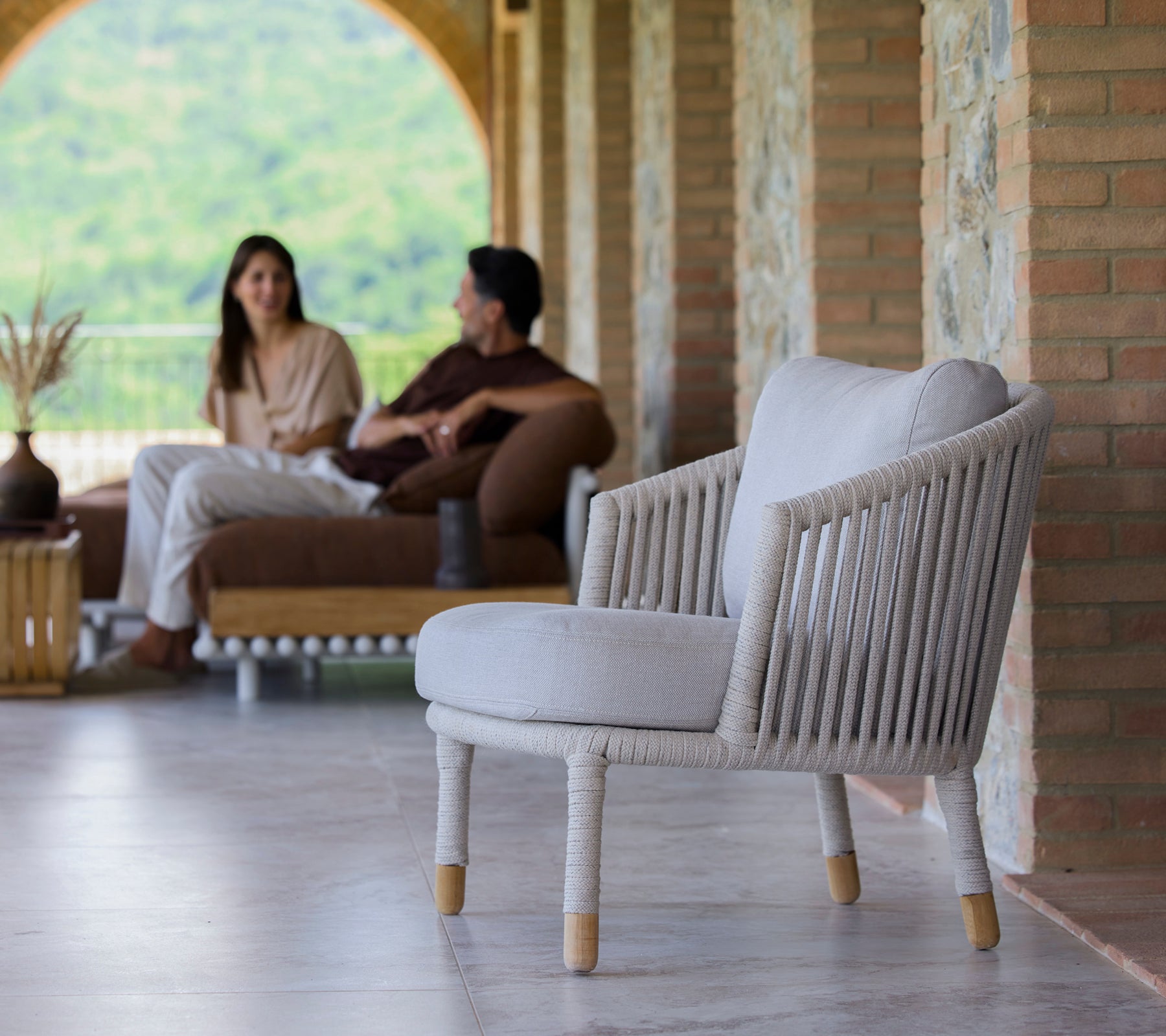Ein sandfarbener Loungesessel mit minimalistischer Design, präsentiert in einer ruhigen Umgebung mit Menschen, die sich unterhalten.