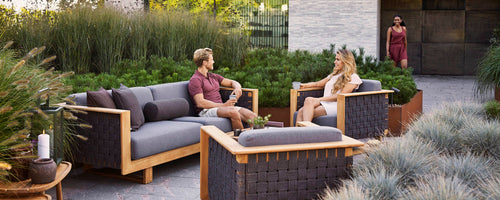 Two people relax on a stylish sofa, surrounded by greenery, engaged in conversation with a third person approaching in the background.