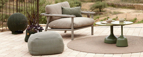 Stilvolles Sitting Area mit einem bequemen Stuhl, einem runden Tisch, einem Pouf und dekorativen Accessoires, die auf einem strukturierten Teppich angeordnet sind.