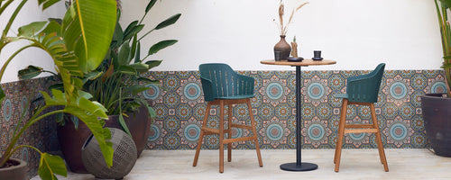 Stylish bar table with two green chairs, surrounded by decorative plants and a patterned backdrop, creating a cozy atmosphere.