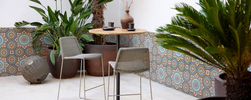 A stylish seating area featuring sleek bar chairs, a wooden table, and lush greenery, complemented by patterned tiles.