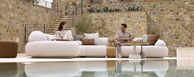 Zwei Personen entspannen sich auf einem stilvollen, modernen Sofa mit Kissen und führen ein Gespräch an einem ruhigen Wasserfall und einer Steinmauer.