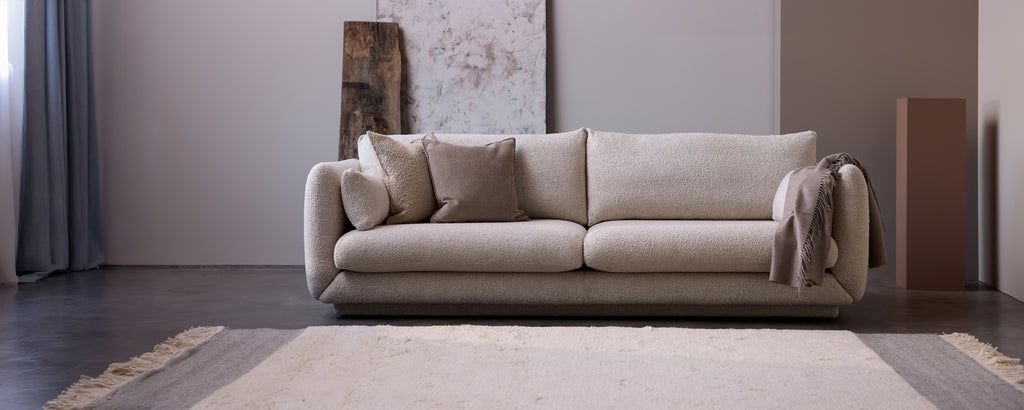 Moderne beigefarbene Sofa mit Kissen und einem grauen Überwurf in einem minimalistischen Wohnzimmer.