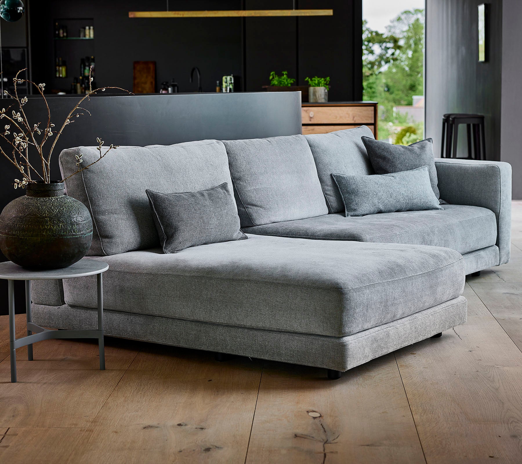 Graues Ecksofa mit strukturiertem Stoff und dekorativen Kissen, begleitet von einem runden Beistelltisch und einer stilvollen Vase.