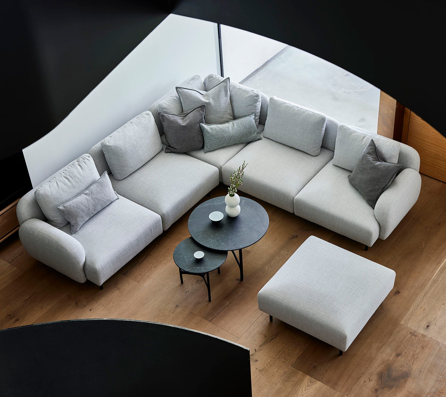 Stylische graue Ecksofa mit dekorativen Kissen, begleitet von einem eleganten runden Couchtisch und einem Hocker auf einem Holzboden.