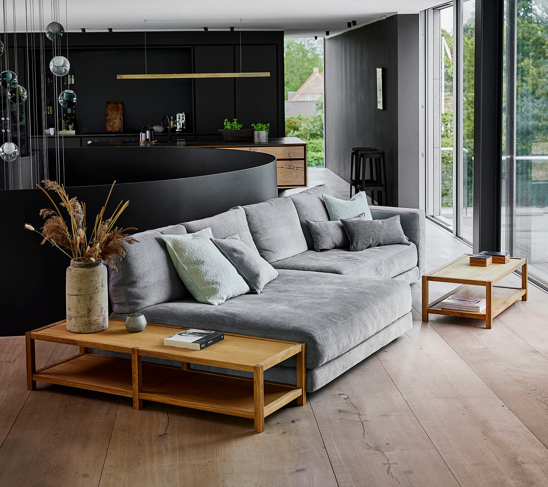 Moderner Wohnraum mit einem grauen Ecksofa, das mit Kissen geschmückt ist, ergänzt durch Holztische und dekorative Pflanzenarrangements.