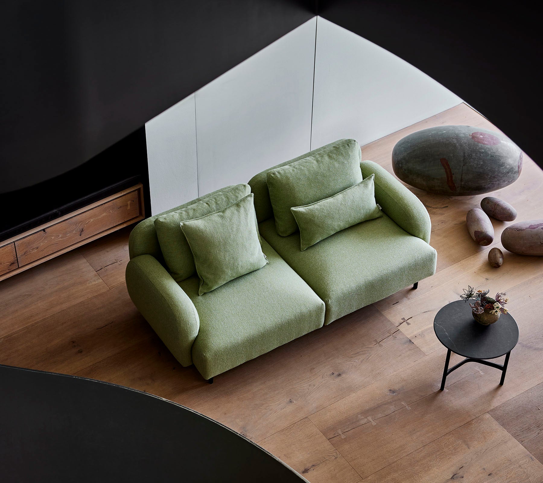 Stylische grüne Couch mit plüschigen Kissen neben einem kleinen runden Tisch, vor einem modernen Hintergrund mit Holzboden und glatten Steinen.