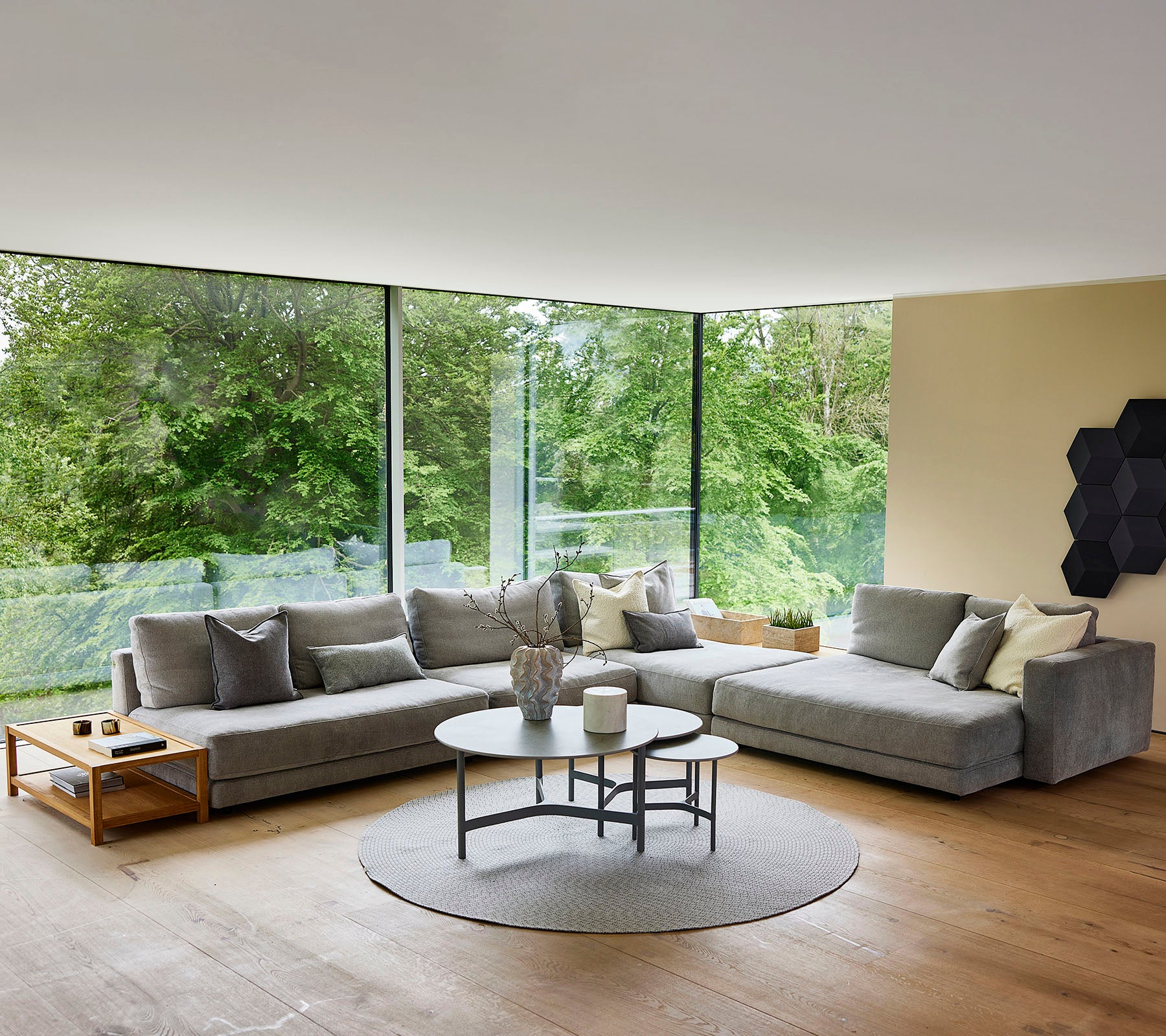 Ein modernes Wohnzimmer mit einem grauen Ecksofa, einem runden Couchtisch und großen Fenstern, die das Tageslicht verstärken.