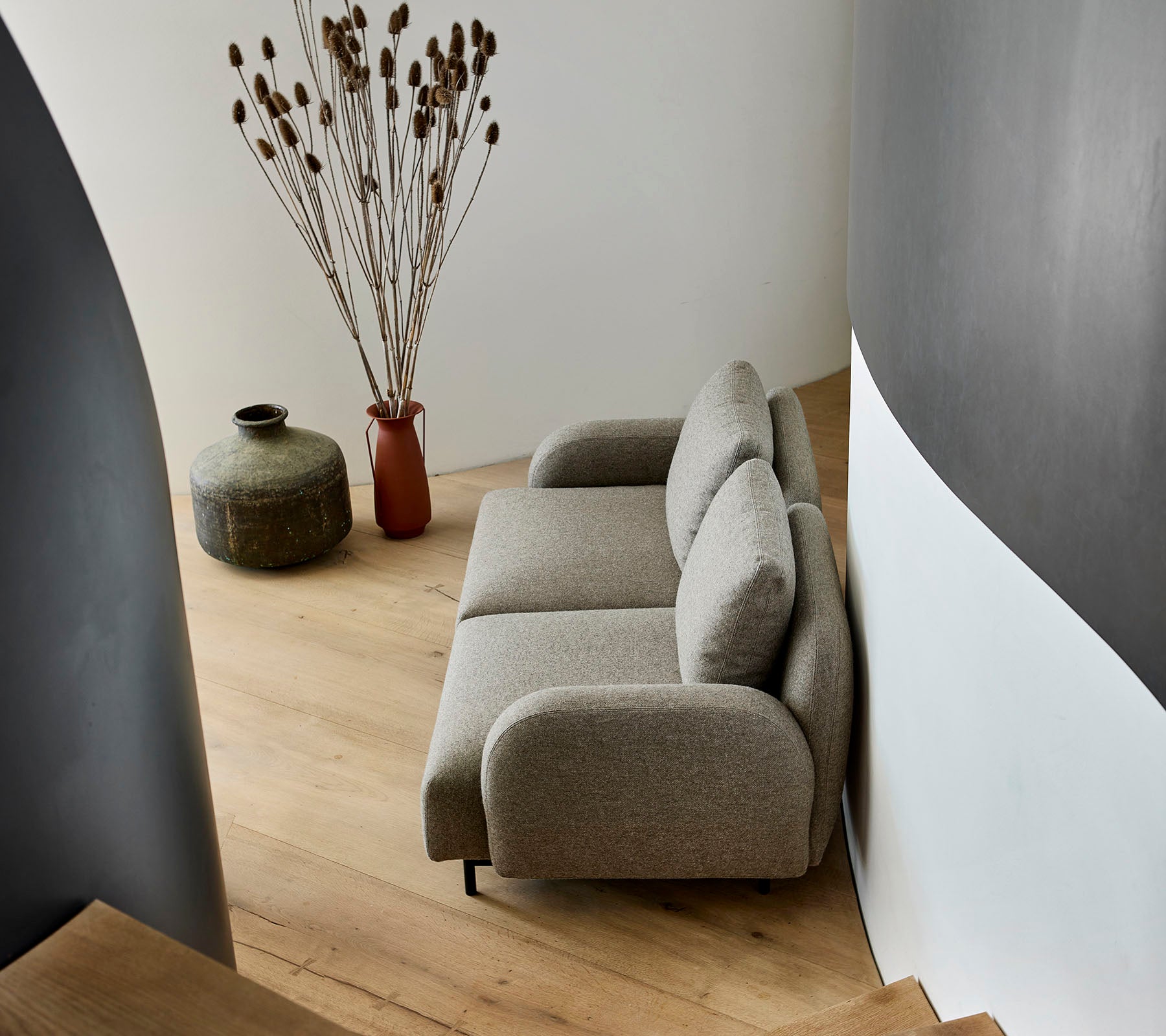 Ein gemütliches beiges Sofa neben einer dekorativen Vase und getrockneten Pflanzen, an einer geschwungenen Wand und einem Holzboden.
