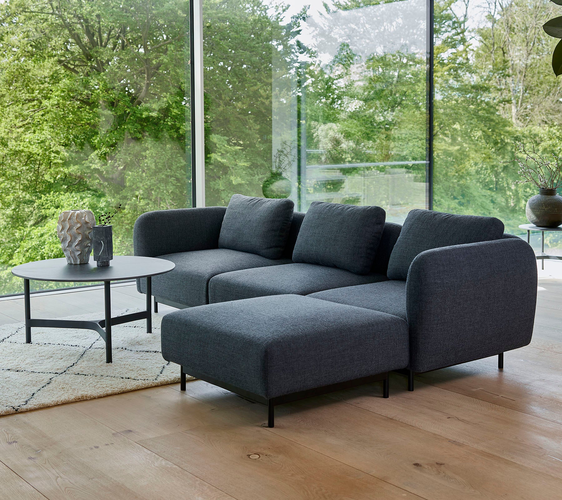 ein modernes graues Ecksofa mit weichen Kissen, kombiniert mit einem eleganten runden Couchtisch auf einem hellen Teppich, umgeben von Grünpflanzen.