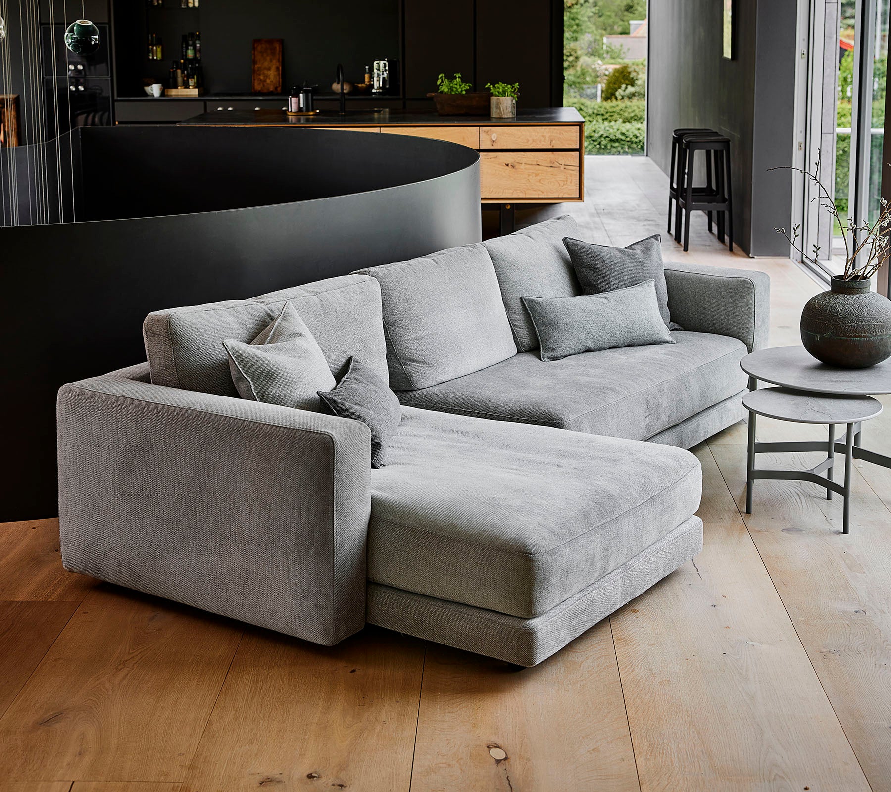 Modernes graues Ecksofa mit mehreren Kissen, begleitet von einem runden Couchtisch und einer dekorativen Vase. Einfaches, elegantes Design.
