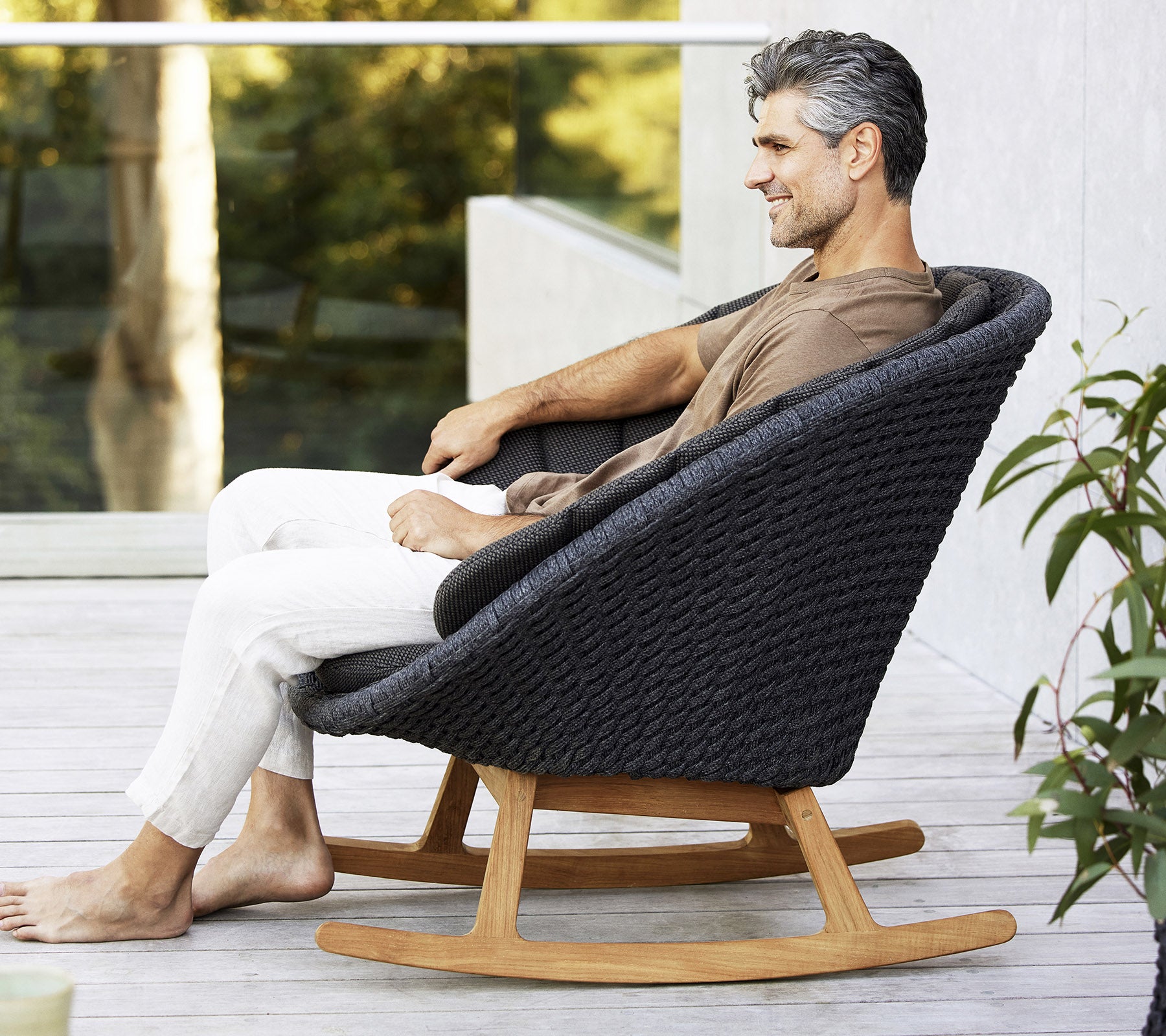 Ein Mann sitzt entspannt in einem stilvollen, modernen Schaukelstuhl mit einem gewobenen Design und einem Fuß aus Naturholz, umgeben von Grünpflanzen.