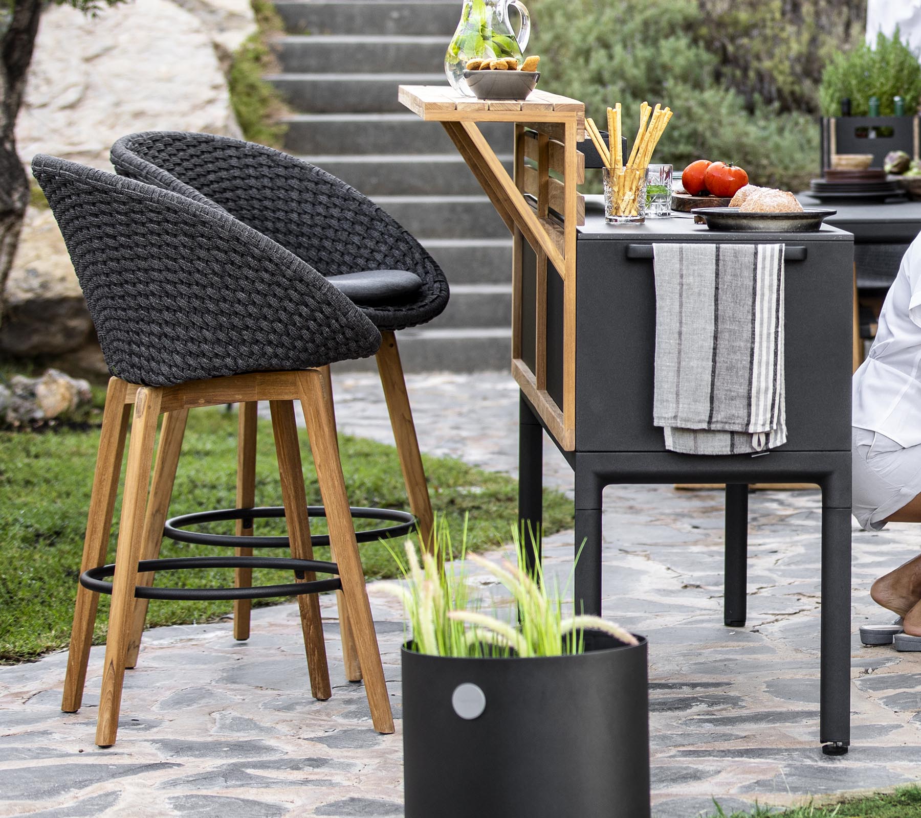 Stylische Außensitzgelegenheiten mit Rattanstühlen, einem modernen Barwagen und frischen Zutaten, die auf einer Arbeitsplatte angeordnet sind, ergänzt durch dekorative Pflanzen.